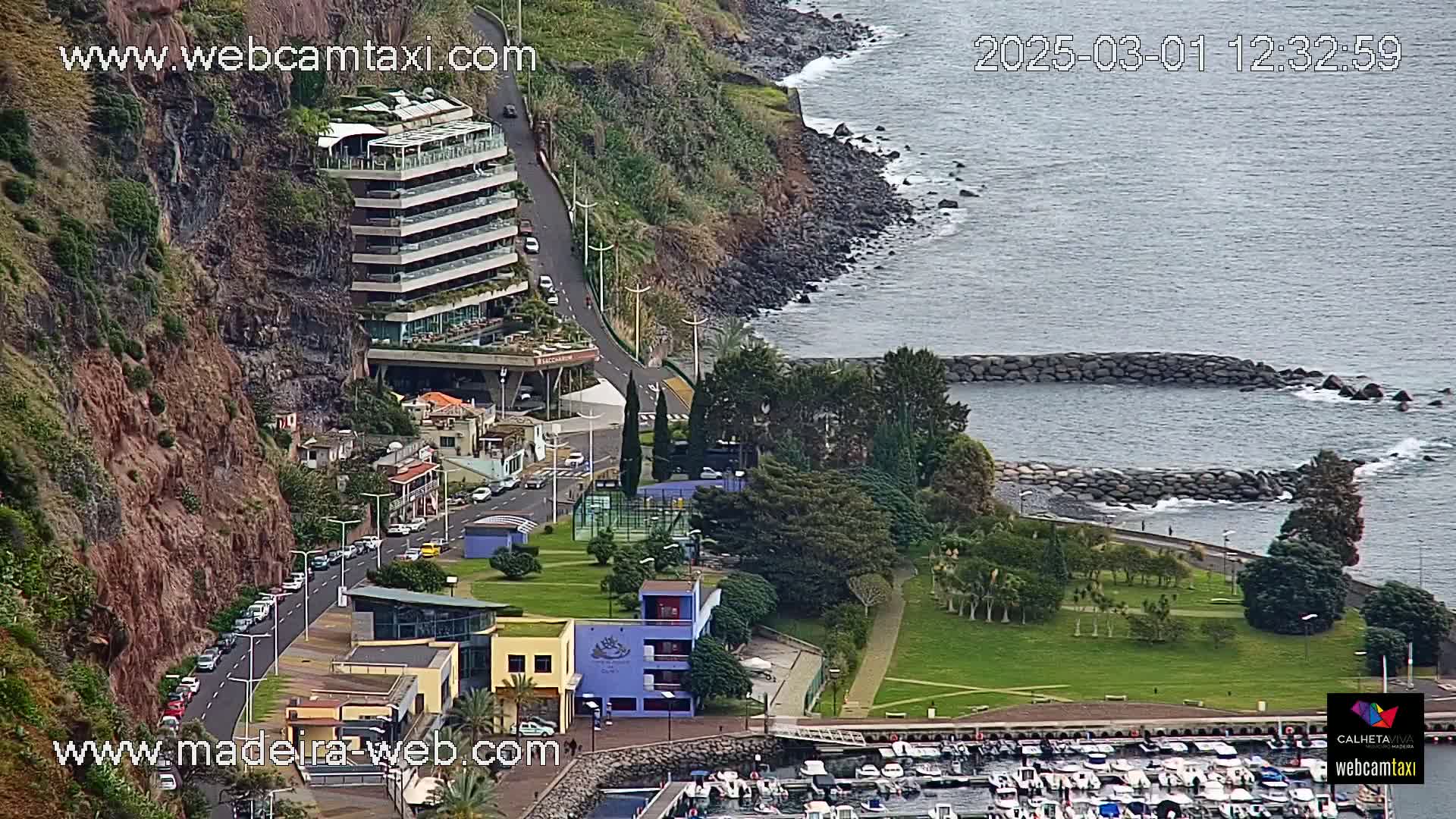 A coastal town with a multi-story building perched on a cliff overlooking a calm ocean with a small marina and green parkland.
