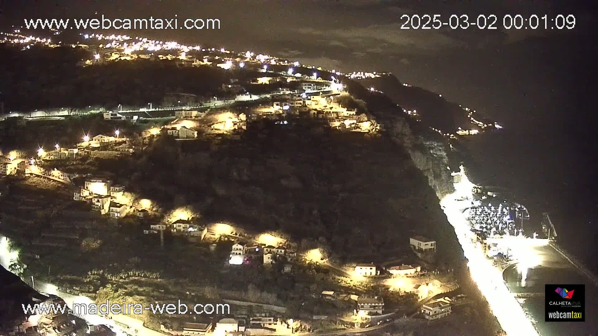A nighttime aerial view of a hillside town illuminated by streetlights, with a road winding down to a coastline under a partly cloudy sky.