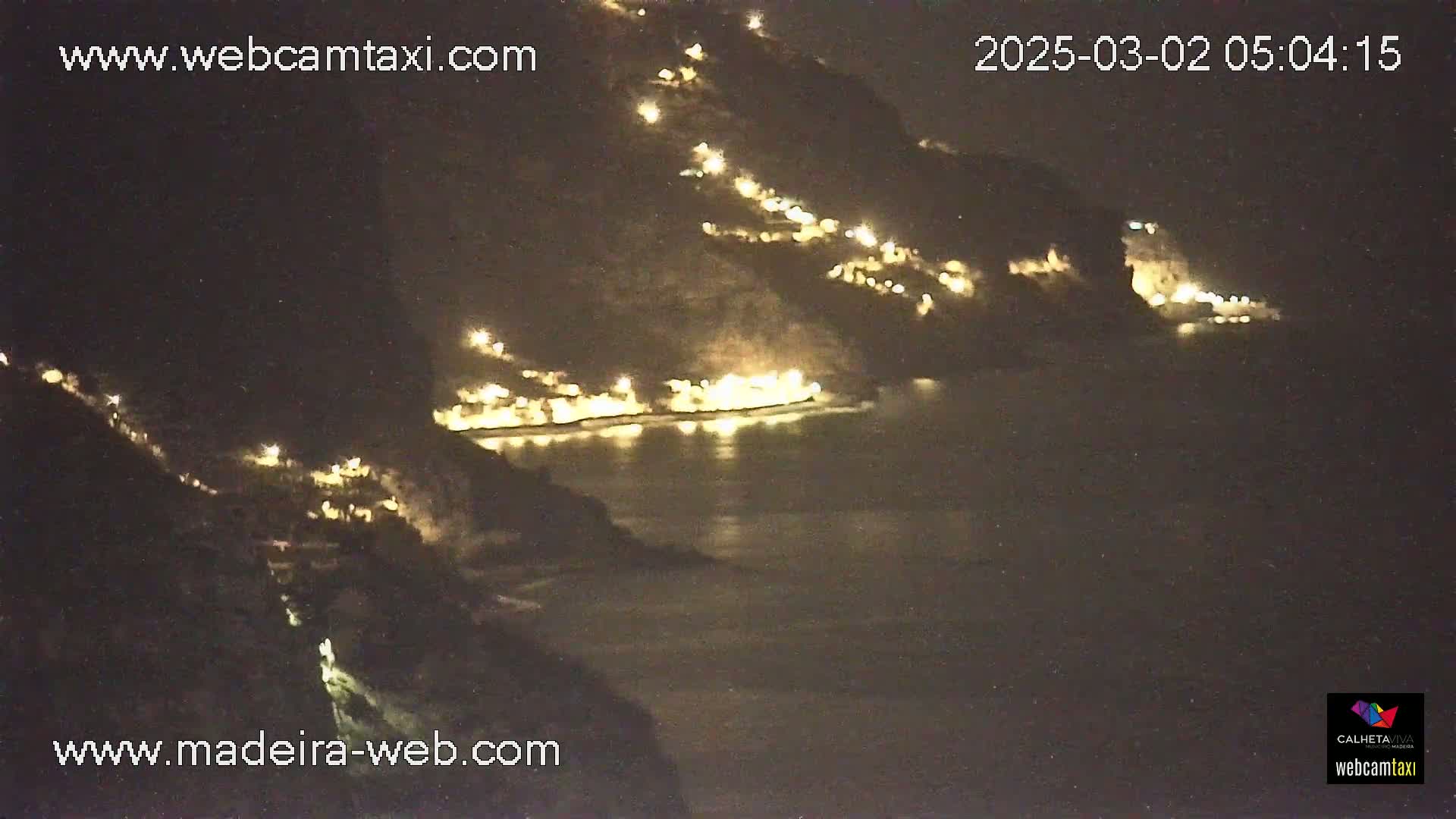 A nighttime coastal scene shows a dark hillside dotted with numerous lights overlooking a calm body of water.