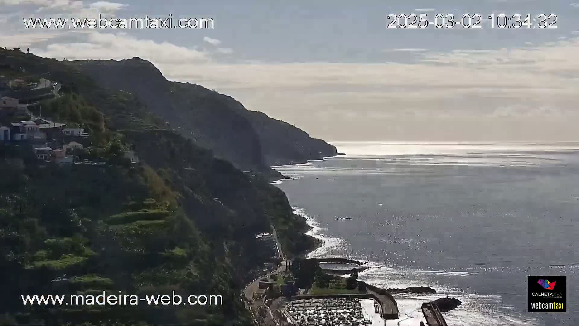 A coastal village nestled on a steep, verdant cliffside overlooks a calm ocean under a partly cloudy sky.