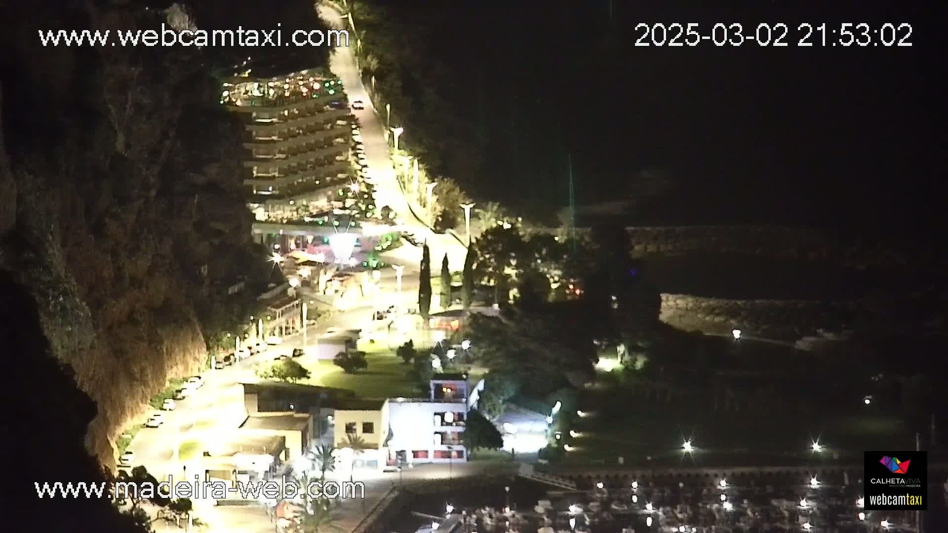 A nighttime, aerial view of a coastal town shows buildings, roads, and a marina illuminated by artificial lights.