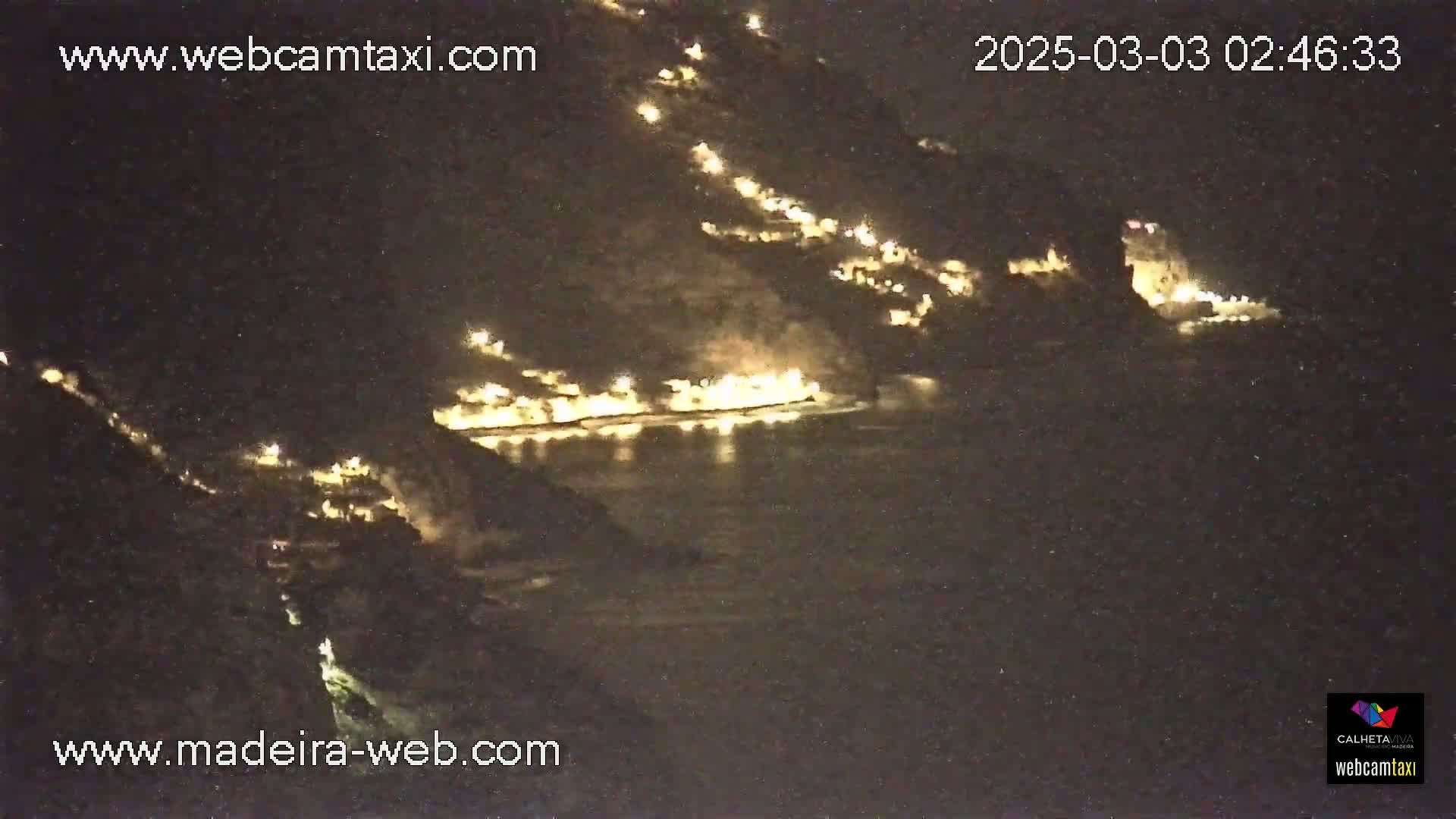 A nighttime, long-range view shows a coastal town's lights nestled between dark hillsides, illuminated against a dark body of water.
