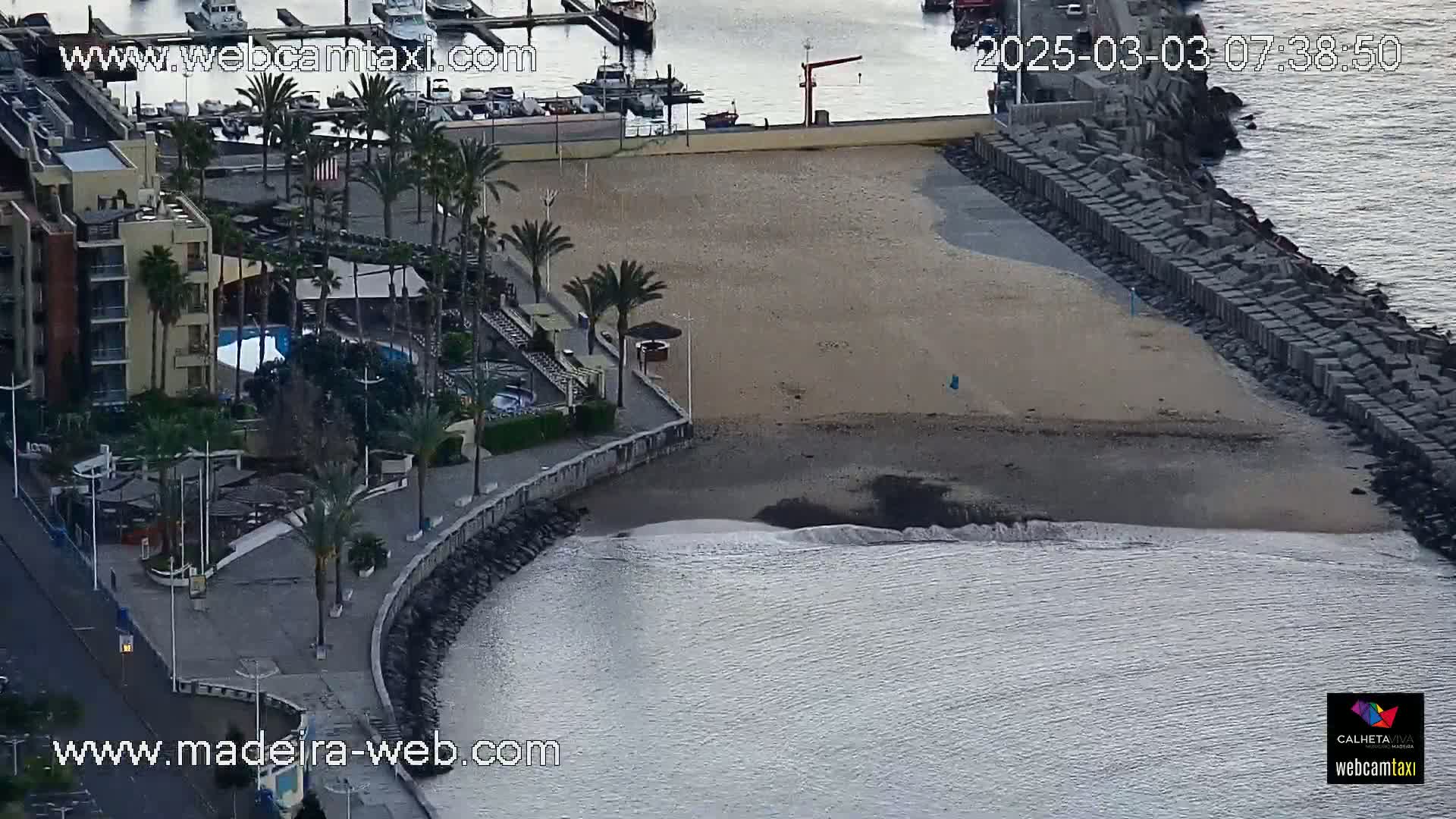 An aerial view shows a sandy beach bordered by a stone wall, a calm ocean, a resort with palm trees, and a marina with boats.