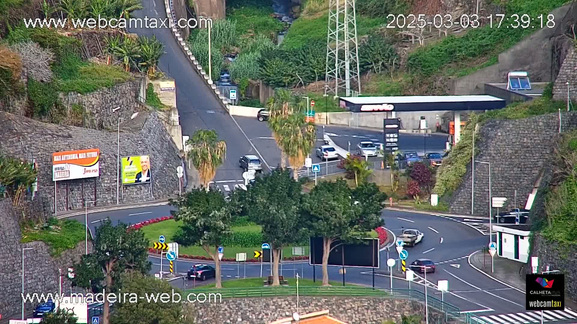 A roundabout with several cars and a gas station is visible, situated on a hillside with lush green vegetation under a partly cloudy sky.