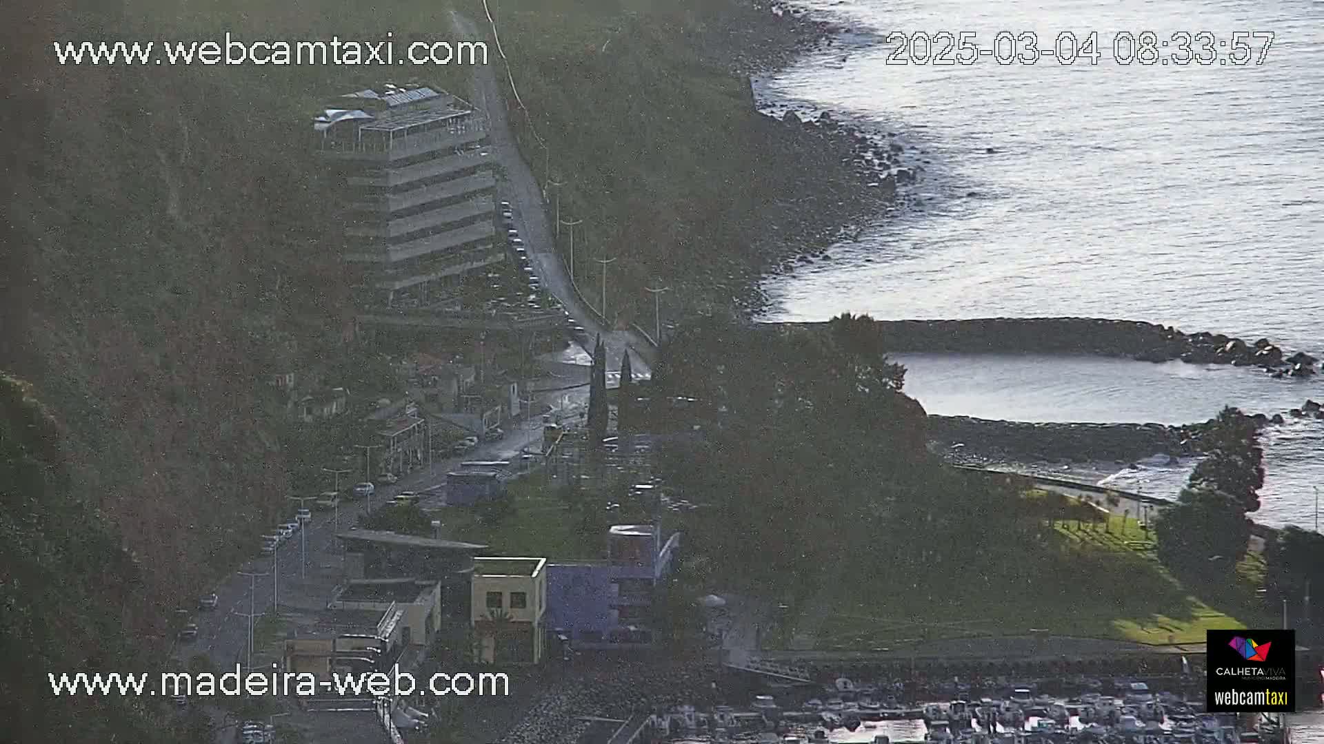 An aerial view shows a coastal town with buildings, a road, and a calm ocean on a partly sunny day.