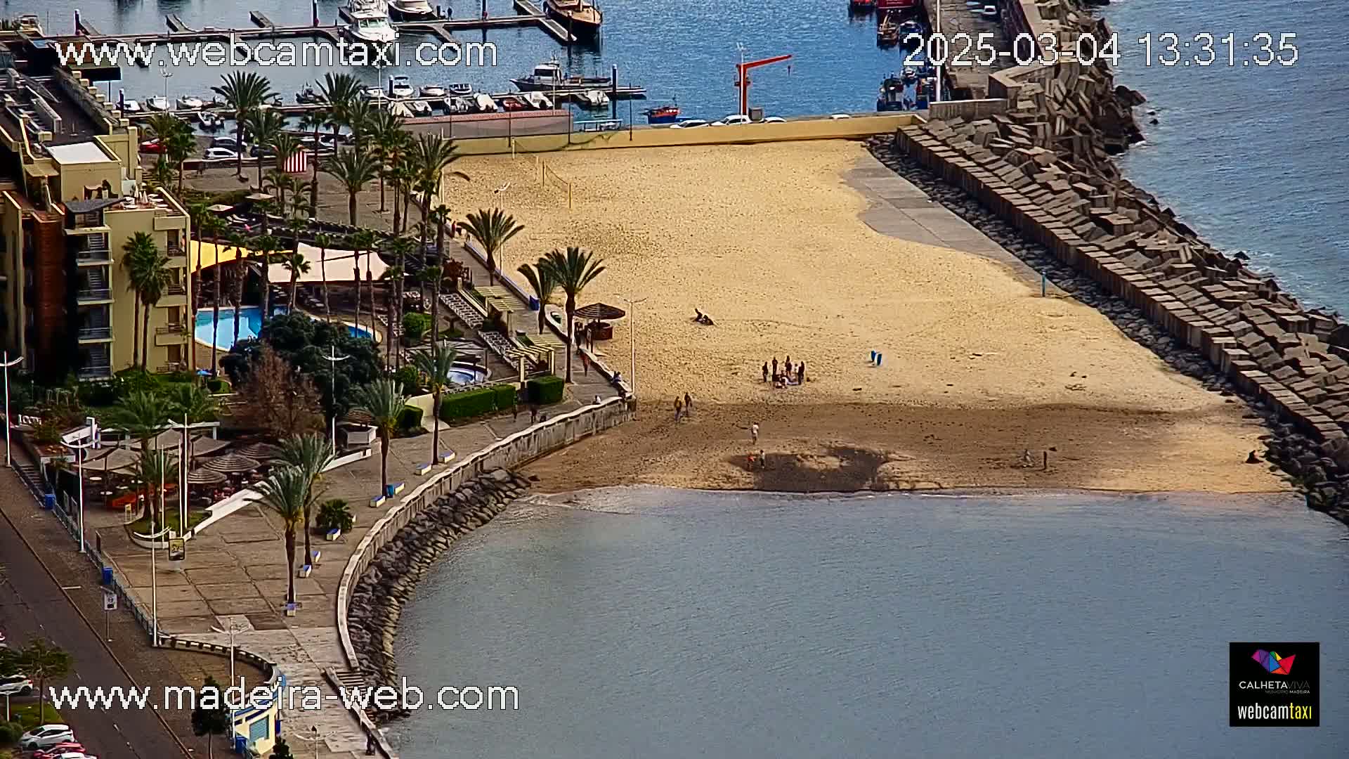 An aerial view shows a sandy beach with a few people on it, next to a marina, a building with a swimming pool, and a rocky breakwater; the water is calm.