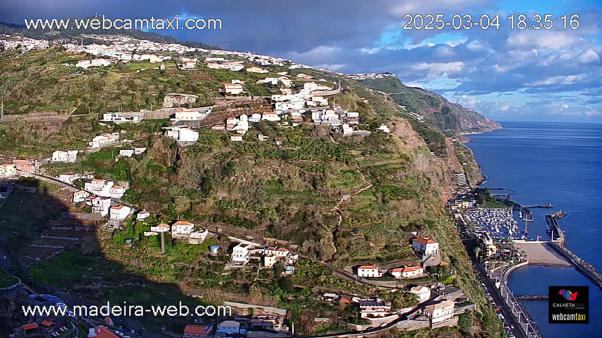 A hillside village clings to a steep cliff overlooking a calm ocean under partly cloudy skies.