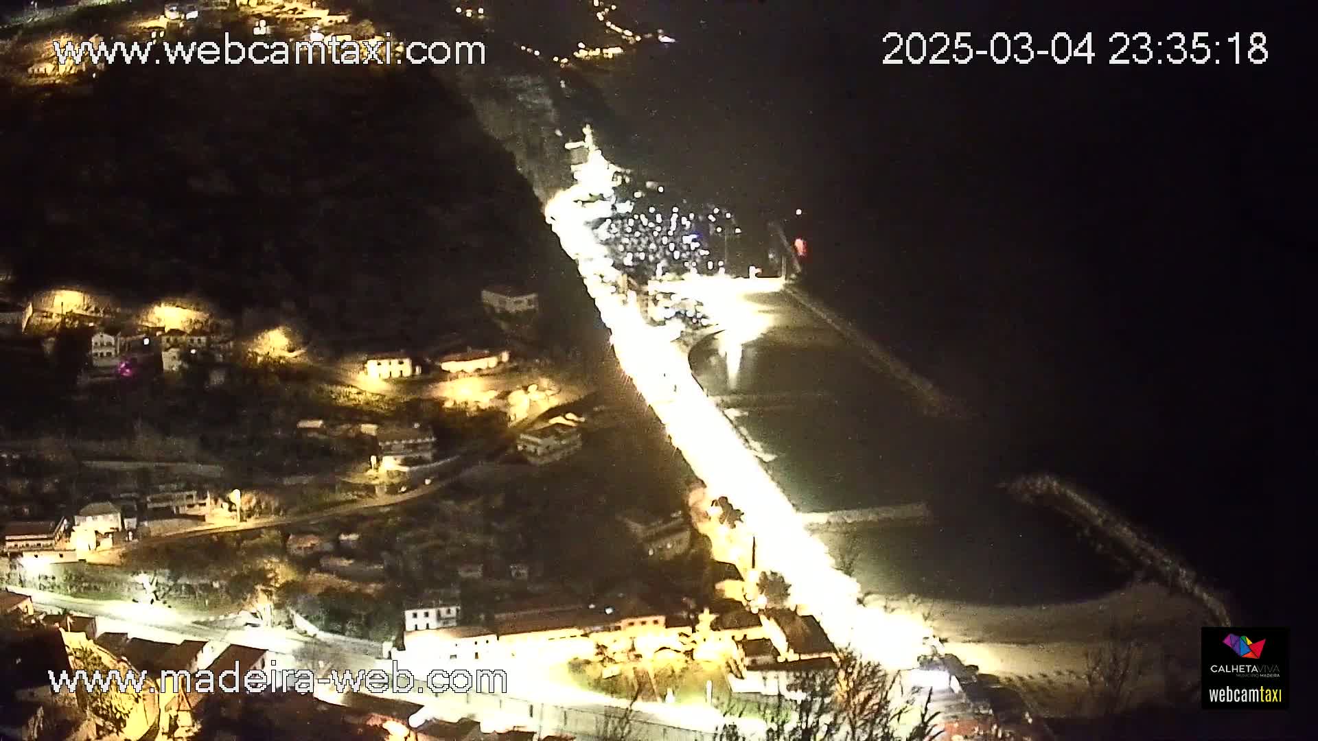 A brightly lit coastal road at night winds through a dark hillside town towards a harbor and body of water.