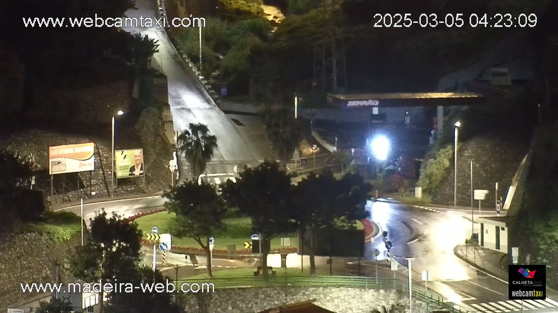 A wet, nighttime scene shows a roadway intersection with a roundabout, surrounded by dark vegetation and buildings under bright streetlights.