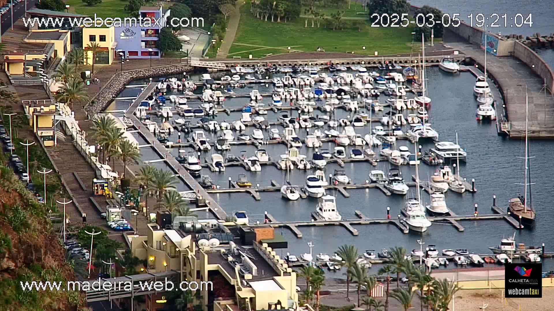 A marina filled with numerous boats and yachts is situated next to a waterfront building complex, under a partly cloudy sky.