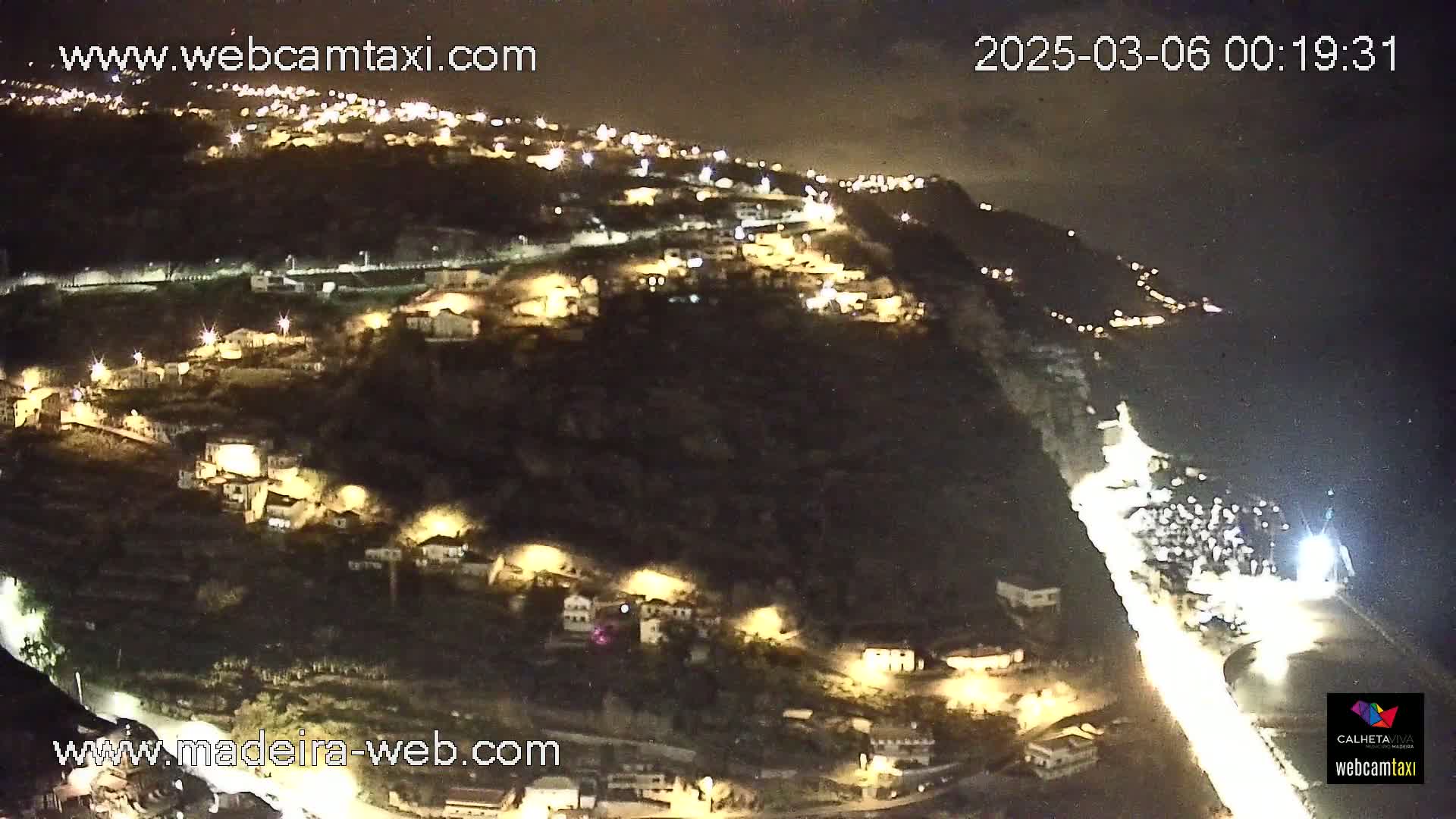 A nighttime, high-angle view shows a hillside village illuminated by lights, with a road winding down to a coastline.