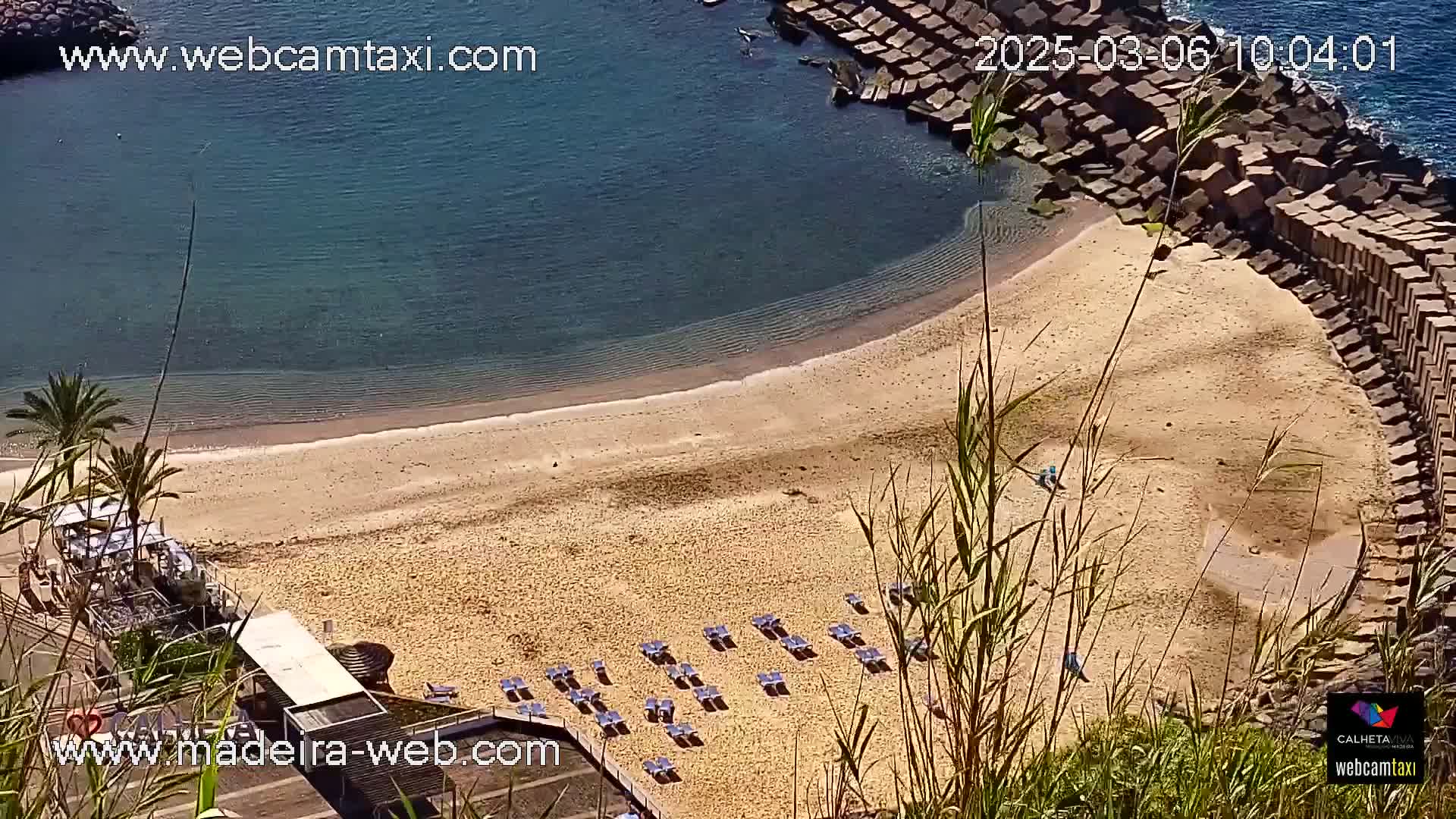 A sandy beach with rows of lounge chairs is nestled between calm, clear blue water and a stone seawall under a sunny sky.