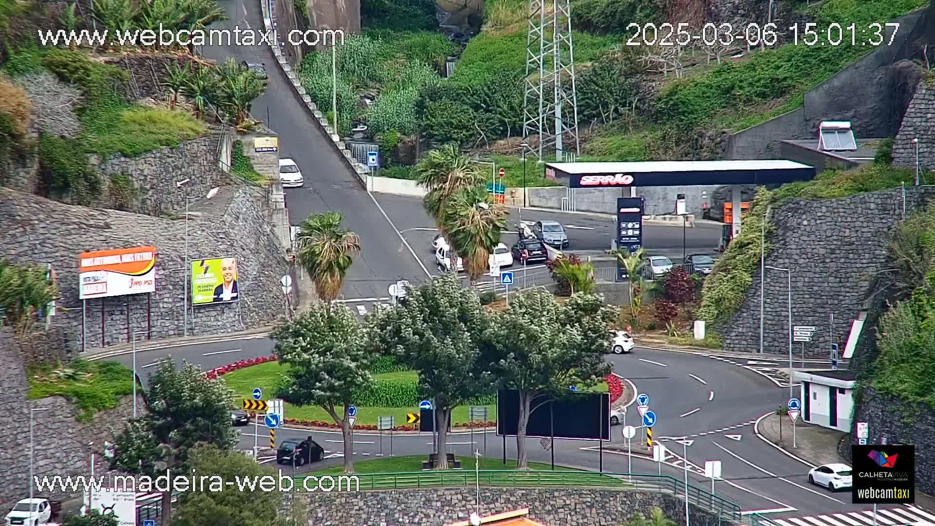 A roundabout with several cars and a gas station is visible from an elevated viewpoint on a partly cloudy day.