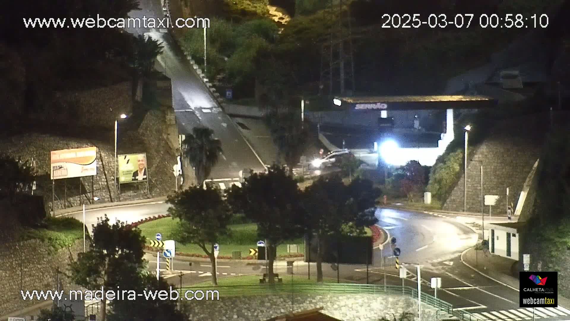 A wet, nighttime roundabout with sparse traffic and surrounding vegetation is illuminated by streetlights.