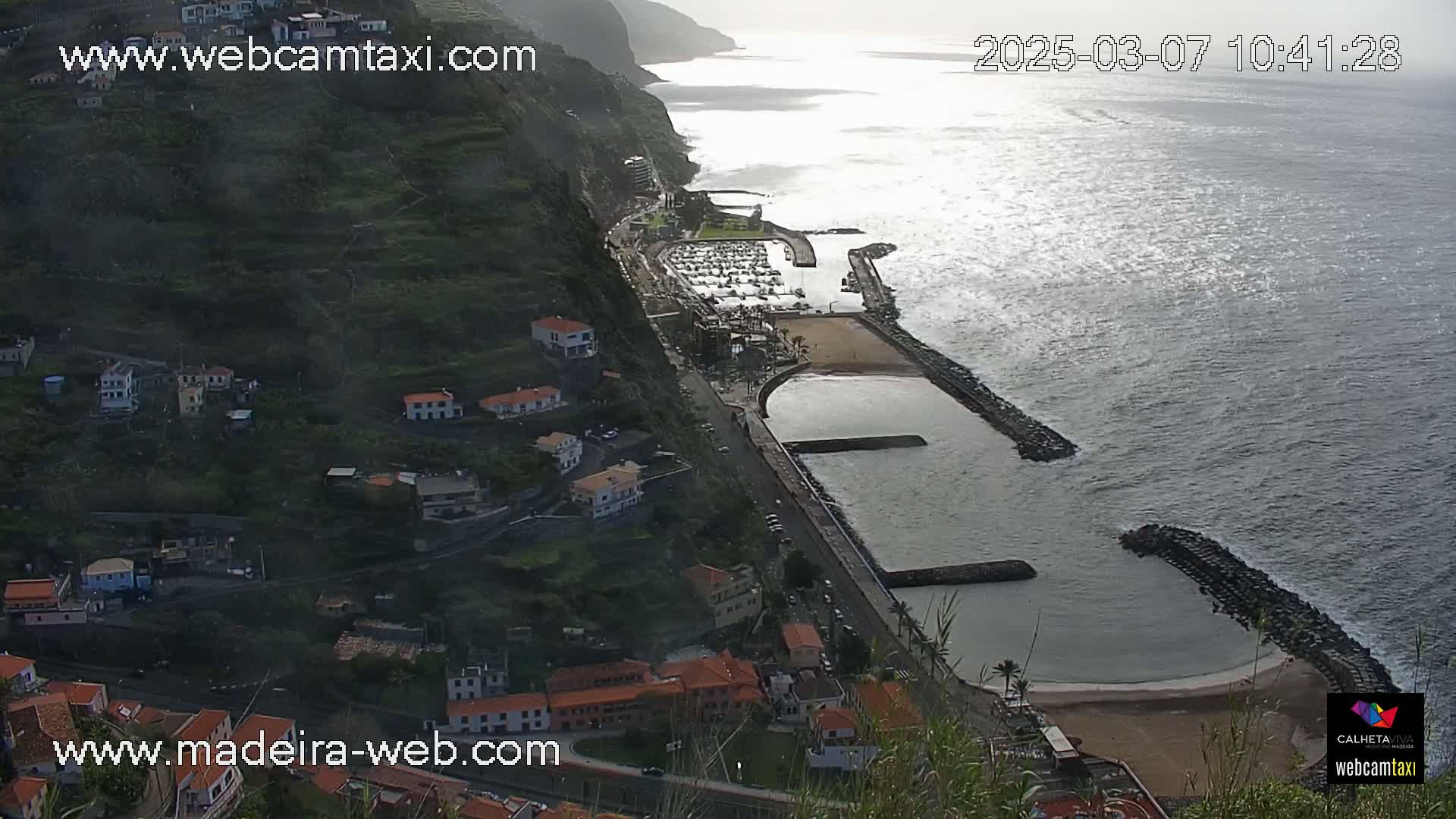 A coastal town nestled on a steep hillside overlooks a calm ocean with a small marina and sandy beach.
