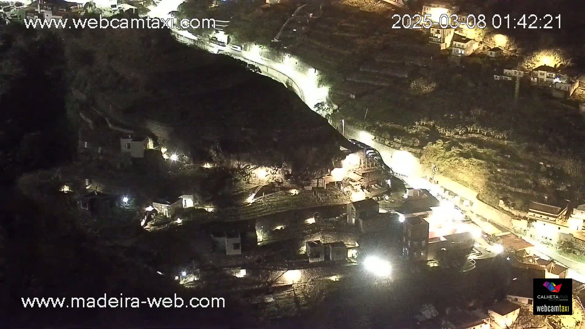 A nighttime aerial view of a hillside village with houses illuminated, a winding road with vehicle lights, and dark, lush vegetation.