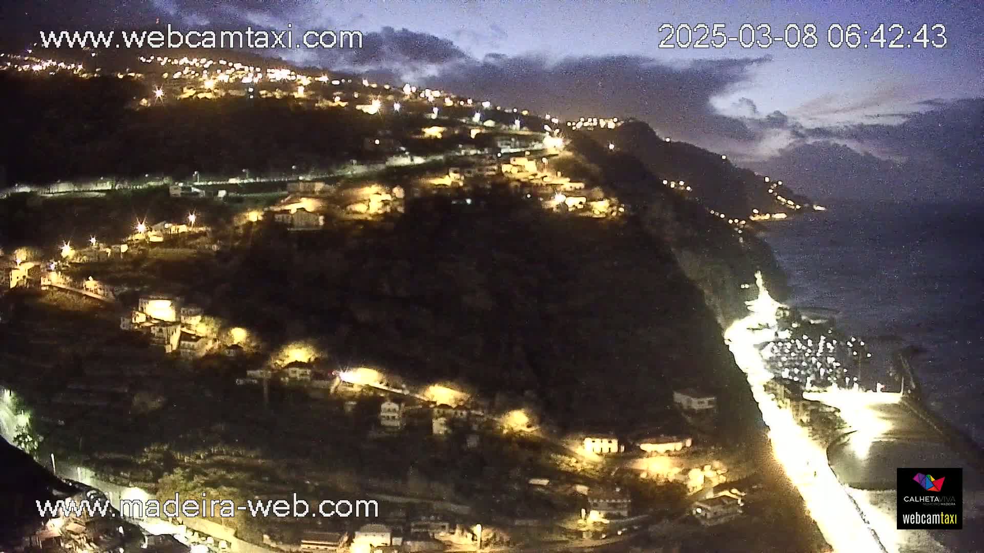 A nighttime coastal village clings to a dark cliffside overlooking a calm ocean under a cloudy sky.
