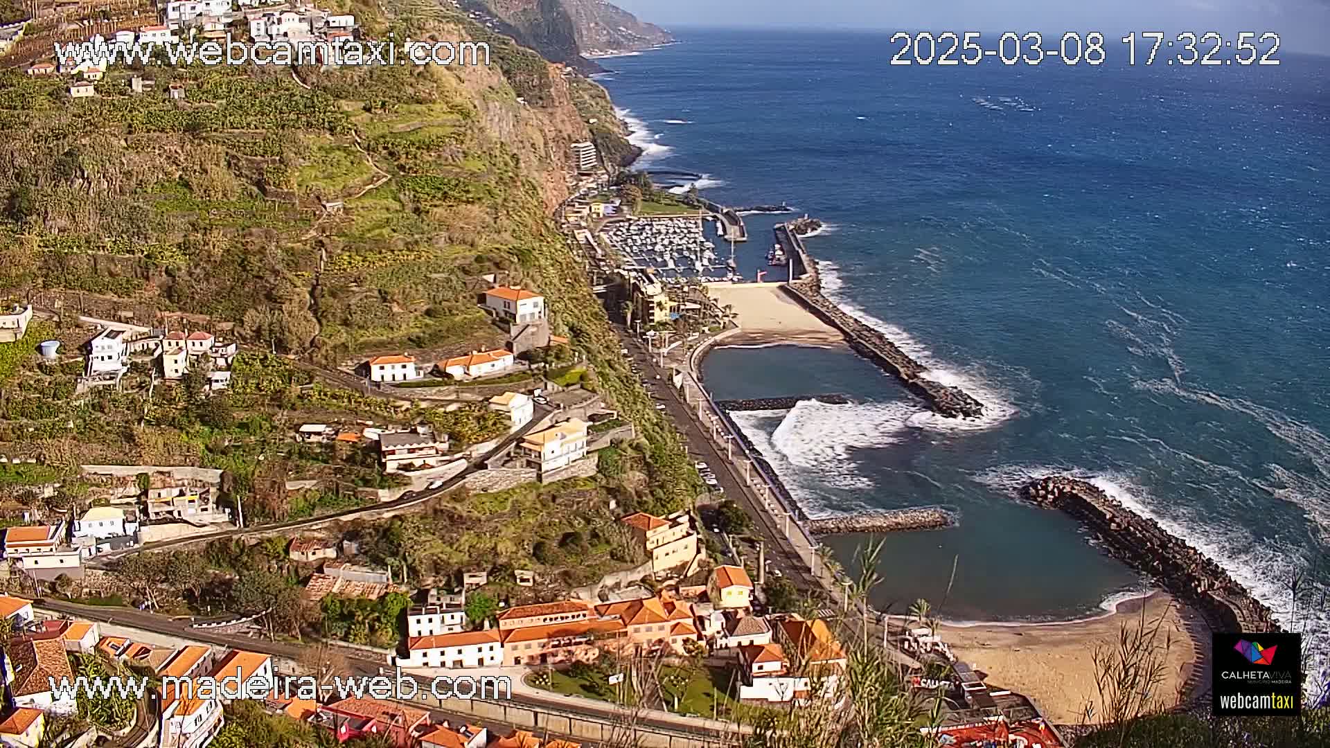 A coastal town nestled on a hillside overlooking a calm ocean with a small marina and sandy beach on a sunny day.