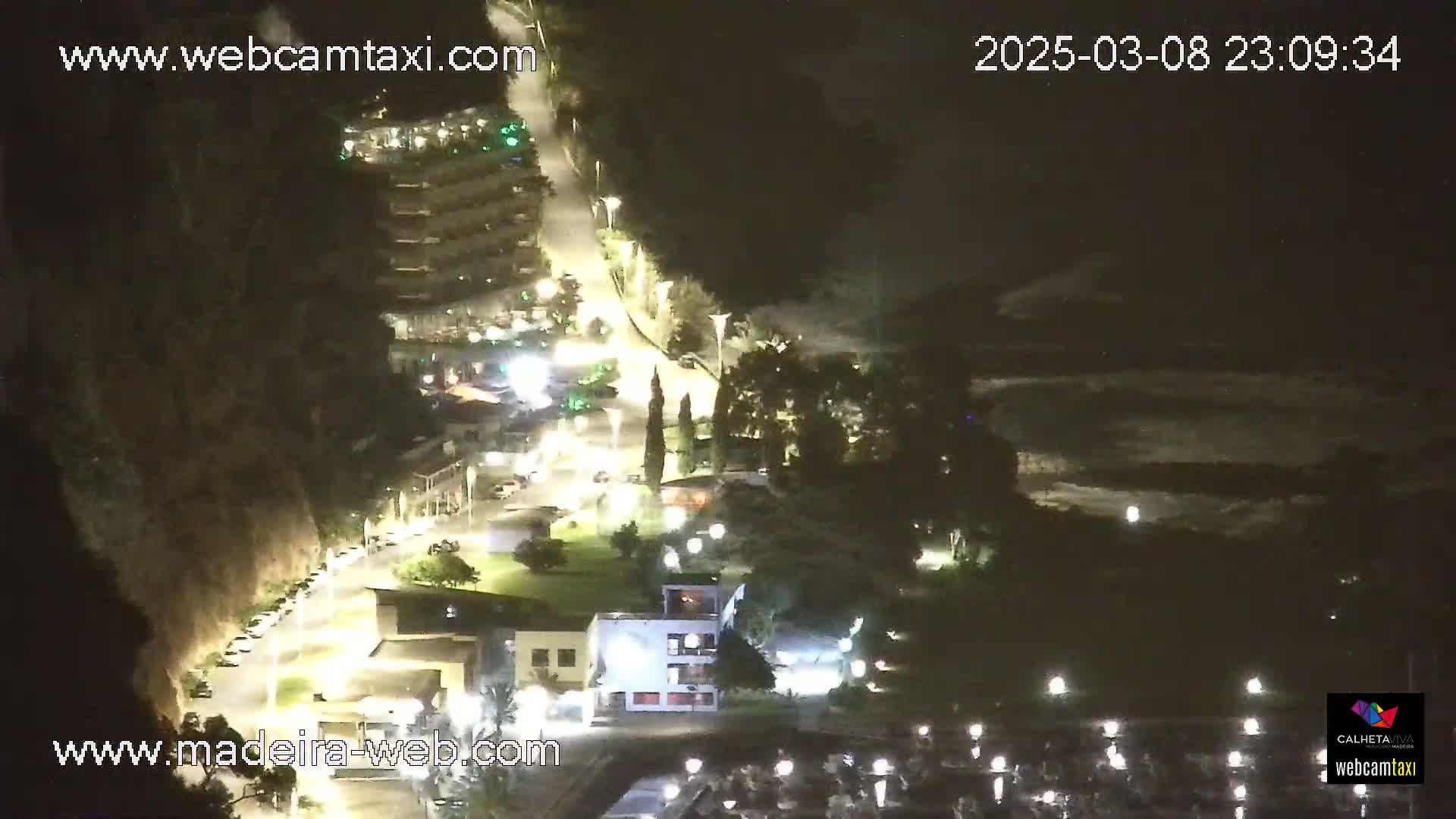 A nighttime aerial view shows a coastal town with buildings, roads, and a marina, under a partly cloudy sky.