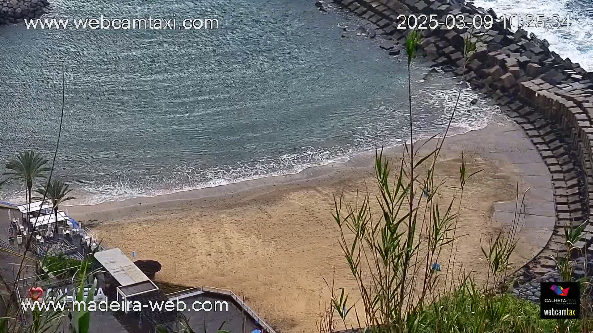 A sandy beach with gentle waves laps against a curved stone wall, overlooking calm ocean water.