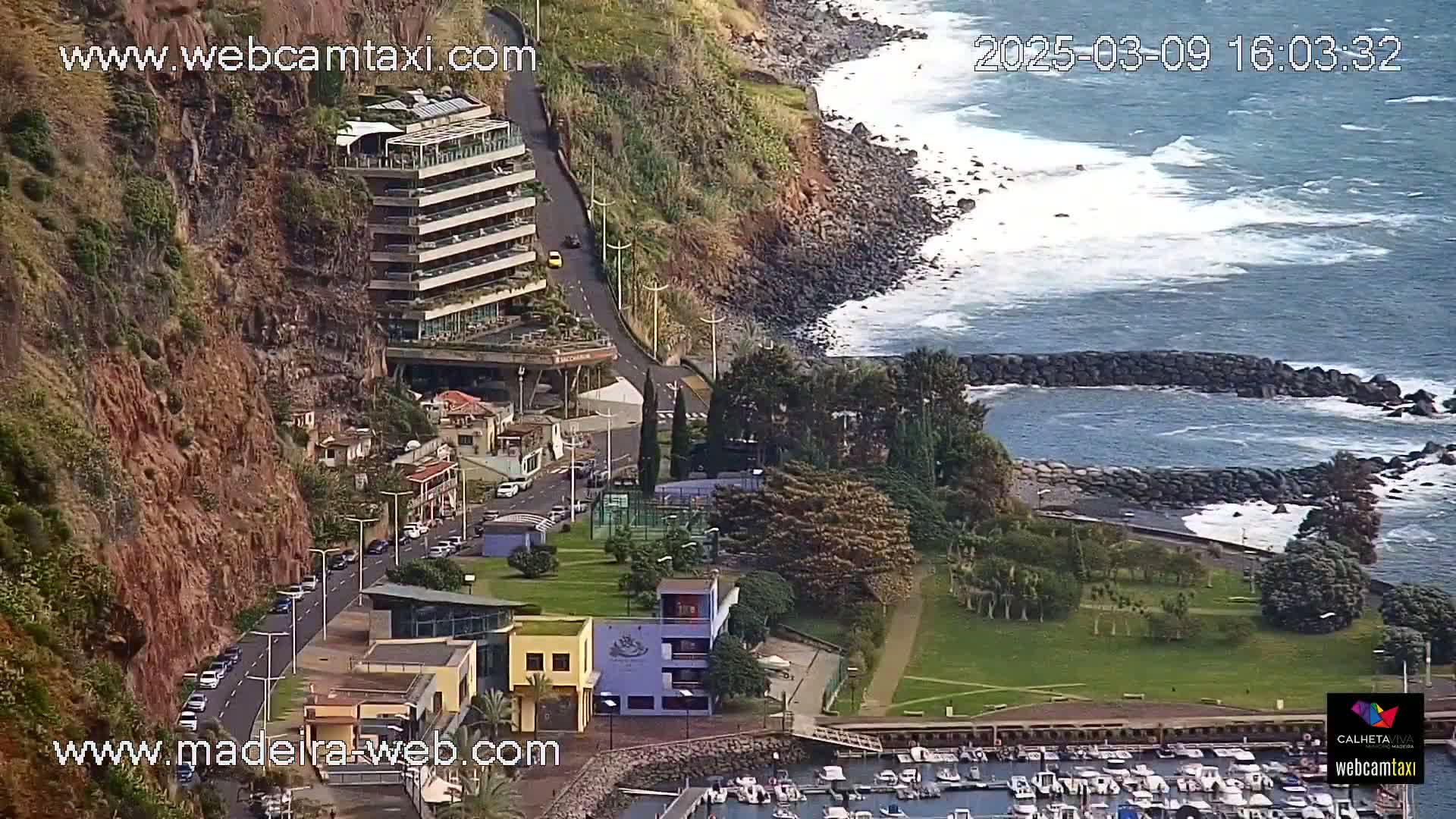 A coastal town with a multi-story building on a cliffside overlooks a marina and ocean with moderate waves.