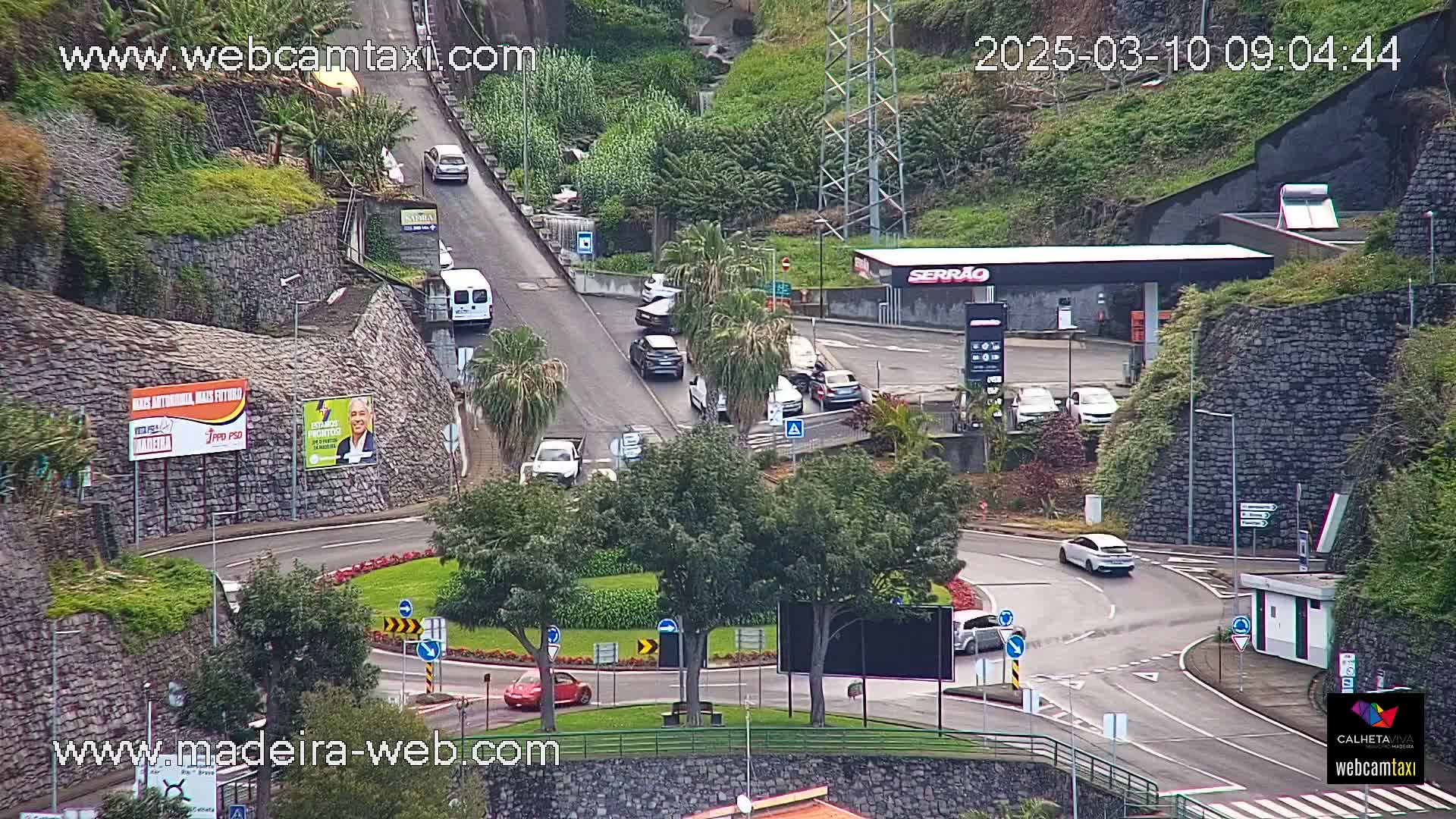 A roundabout with several cars and a gas station is situated on a hillside overlooking a winding road, under overcast conditions.