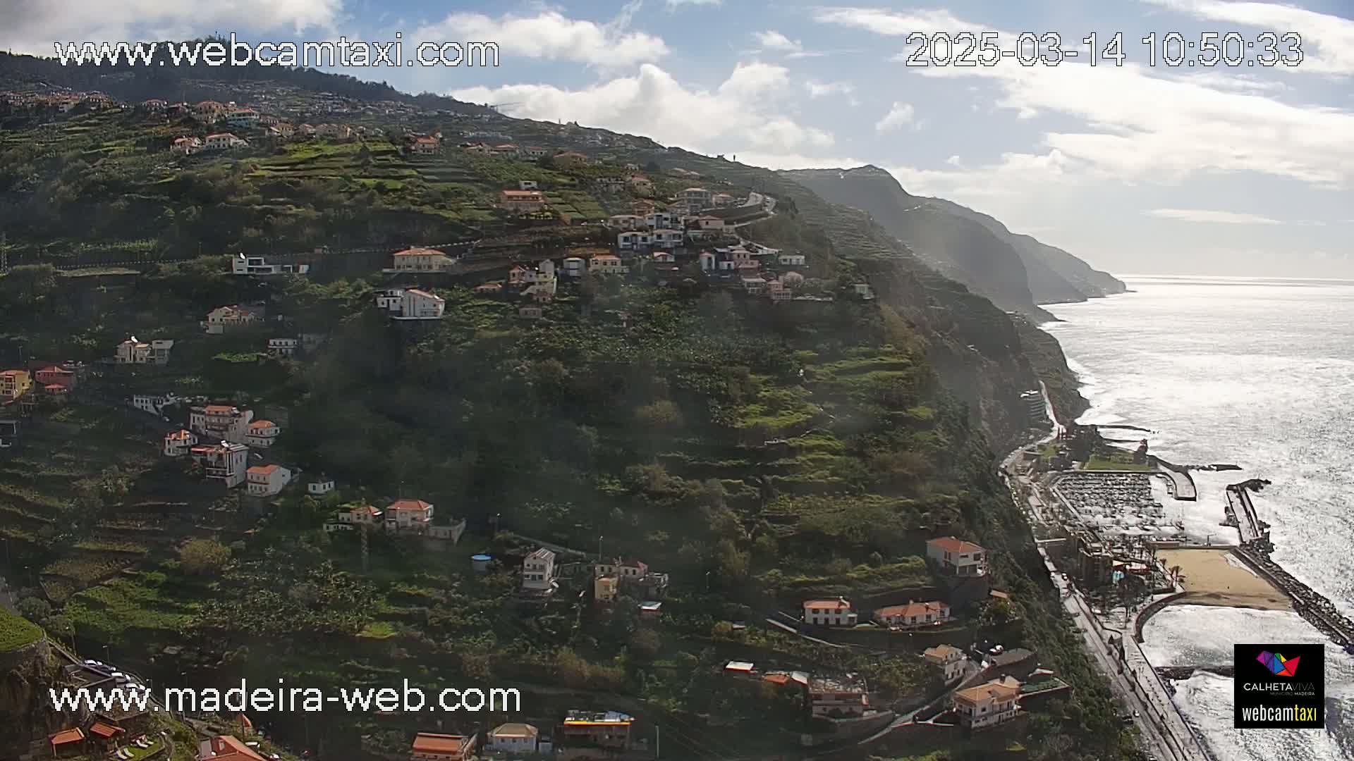 A hillside village clings to a steep, terraced cliff overlooking a calm ocean under a partly cloudy sky.