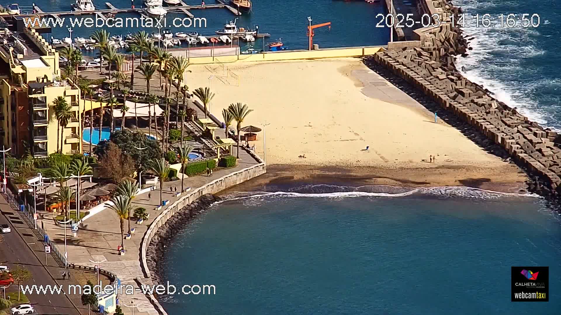 An aerial view shows a sandy beach bordered by a stone seawall, a resort with a pool, palm trees, and calm ocean waves under sunny skies.