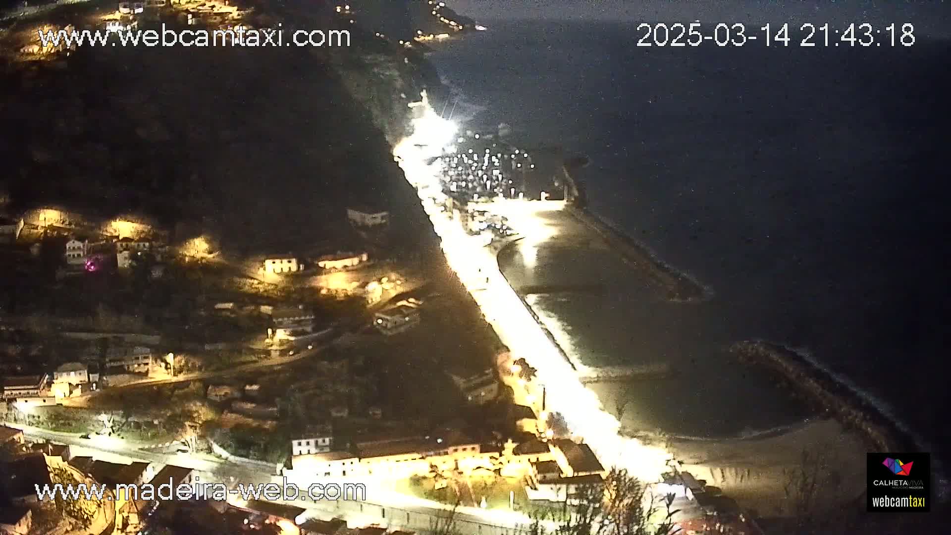 A nighttime, high-angle view shows a coastal town with brightly lit roads and buildings next to a dark ocean with gentle waves breaking against a seawall.