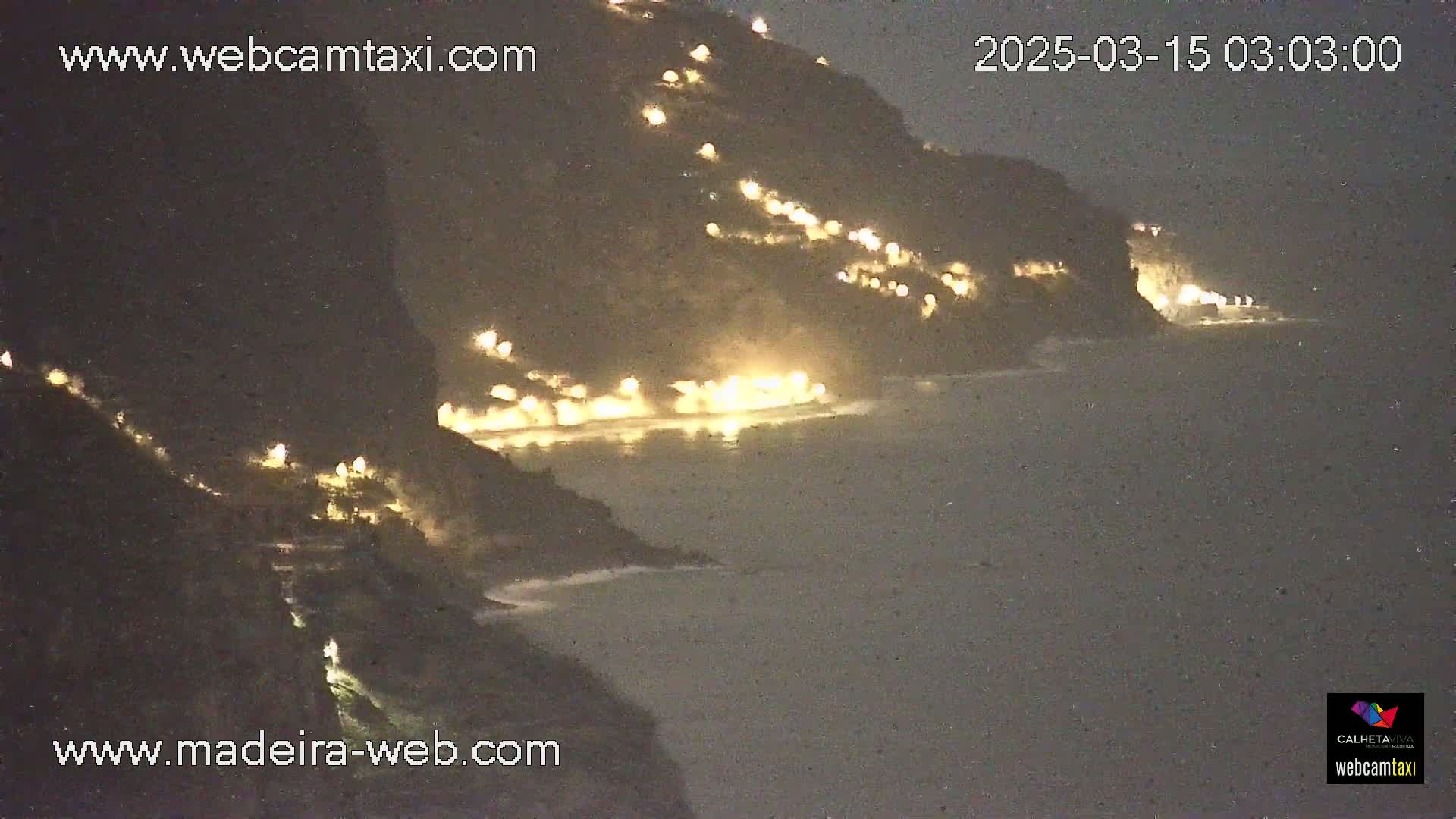 A nighttime coastal scene shows a dark cliffside with scattered lights illuminating buildings, overlooking a dark ocean with gentle waves.