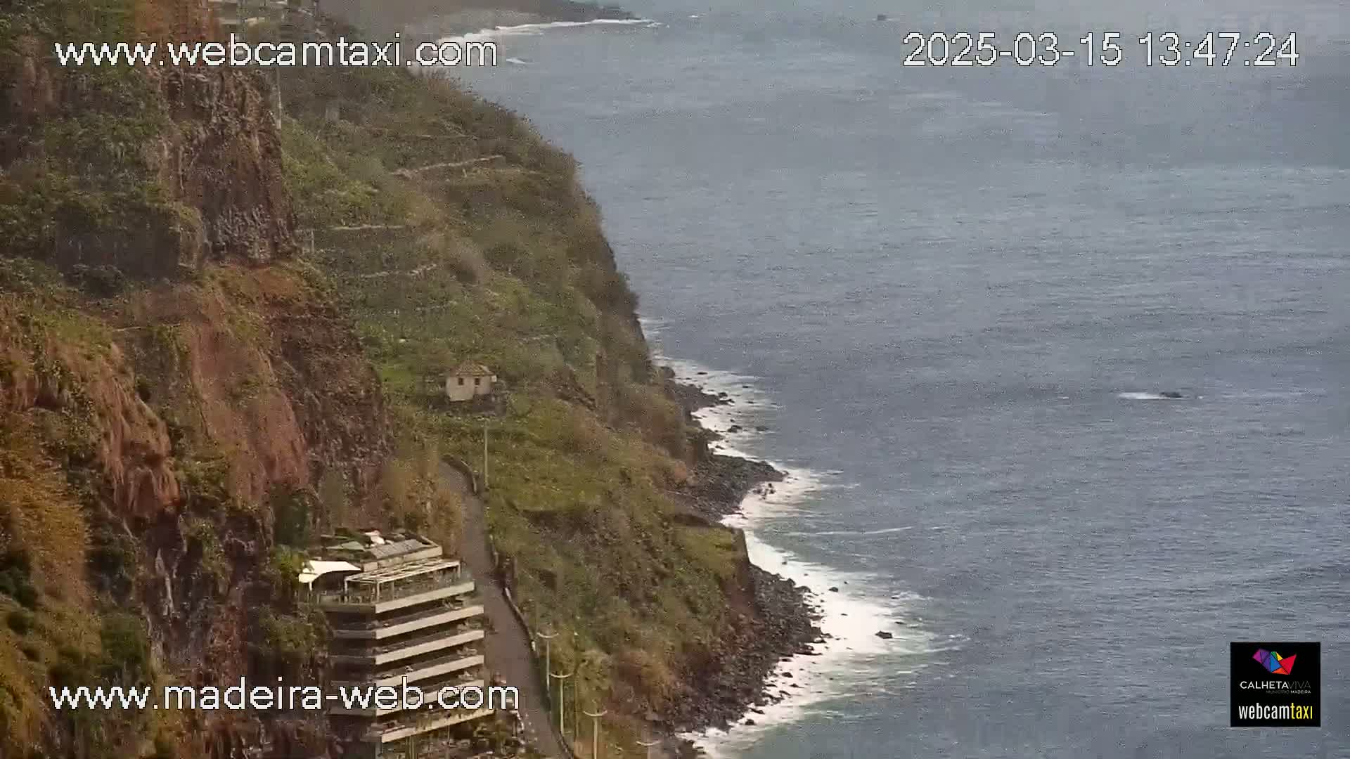 A rocky cliffside with a building and road overlooks a dark-blue ocean with small waves on a cloudy day.