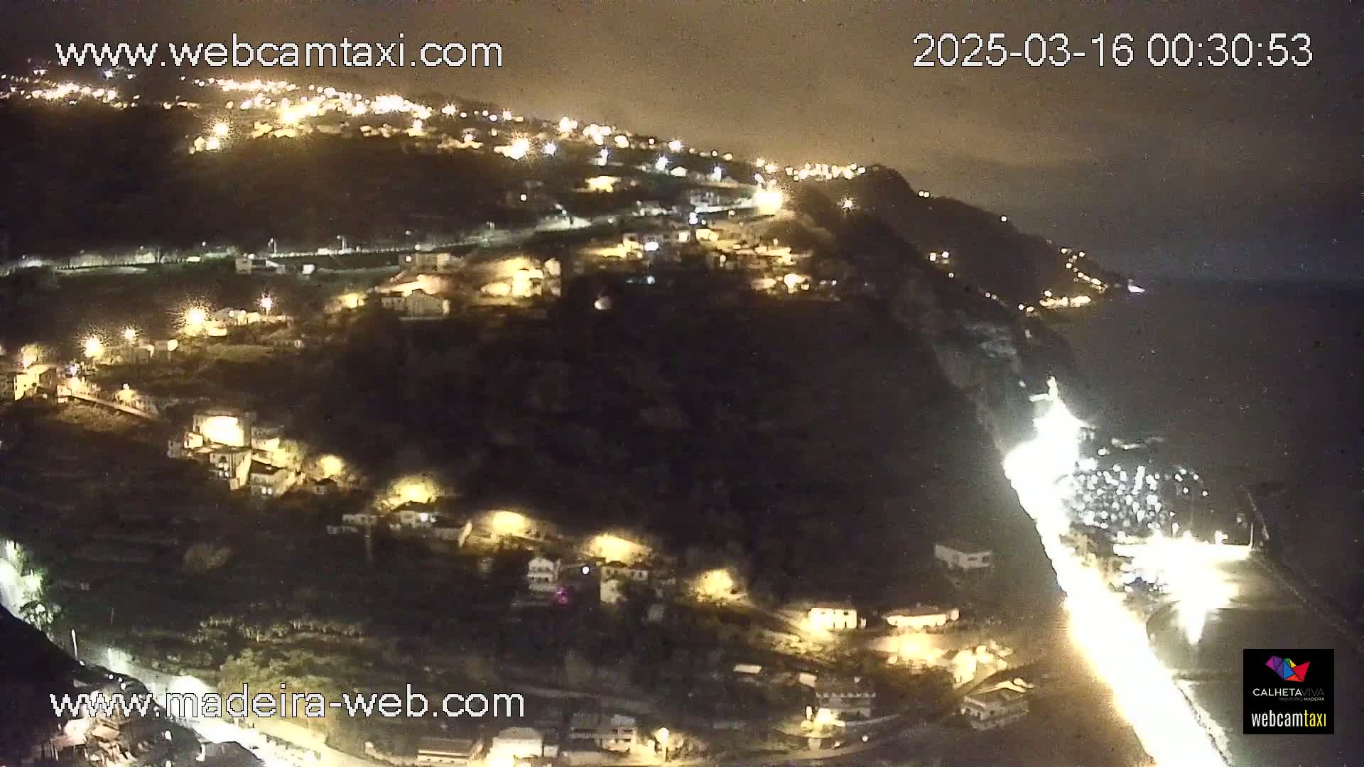A nighttime, high-angle view shows a coastal town illuminated by streetlights, with a road winding along the cliffside toward the ocean.