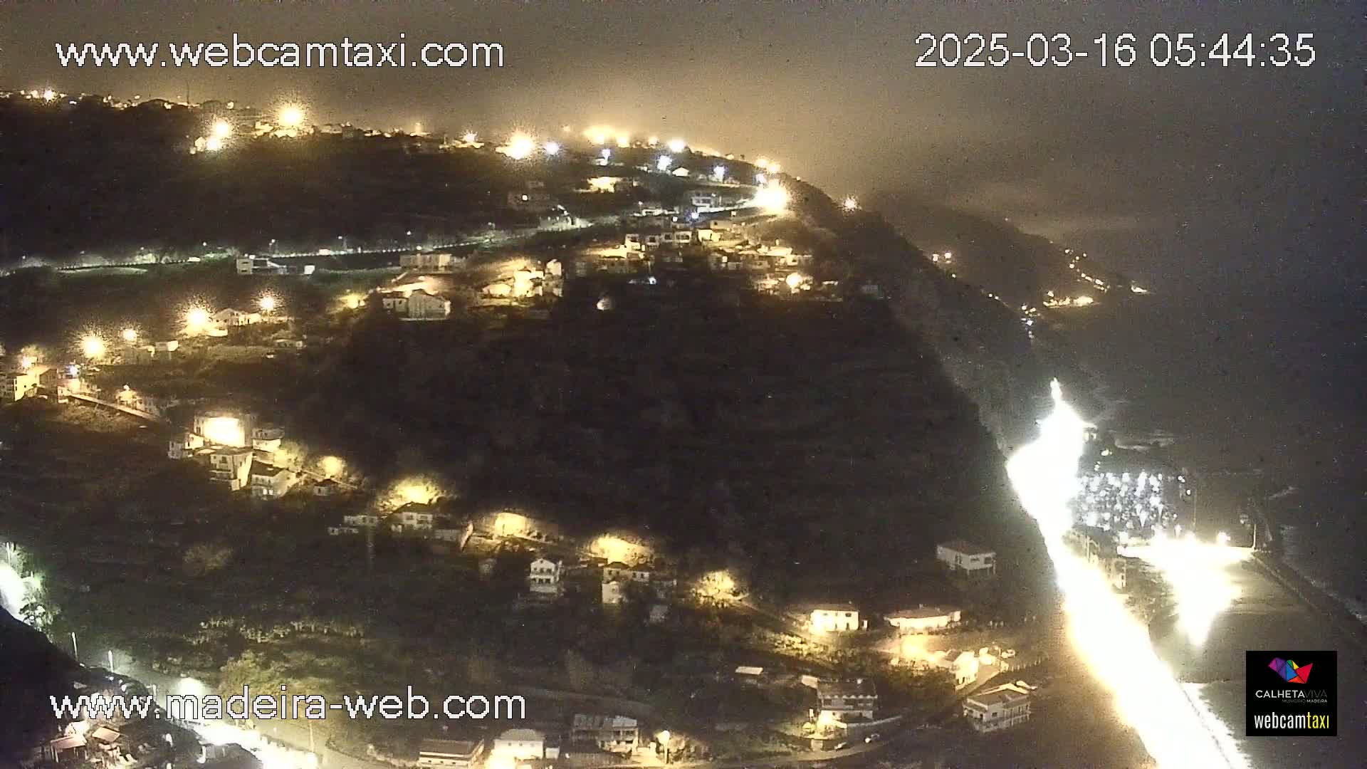 A nighttime, hazy view of a hillside town illuminated by streetlights, with a bright roadway winding down to a coastline.