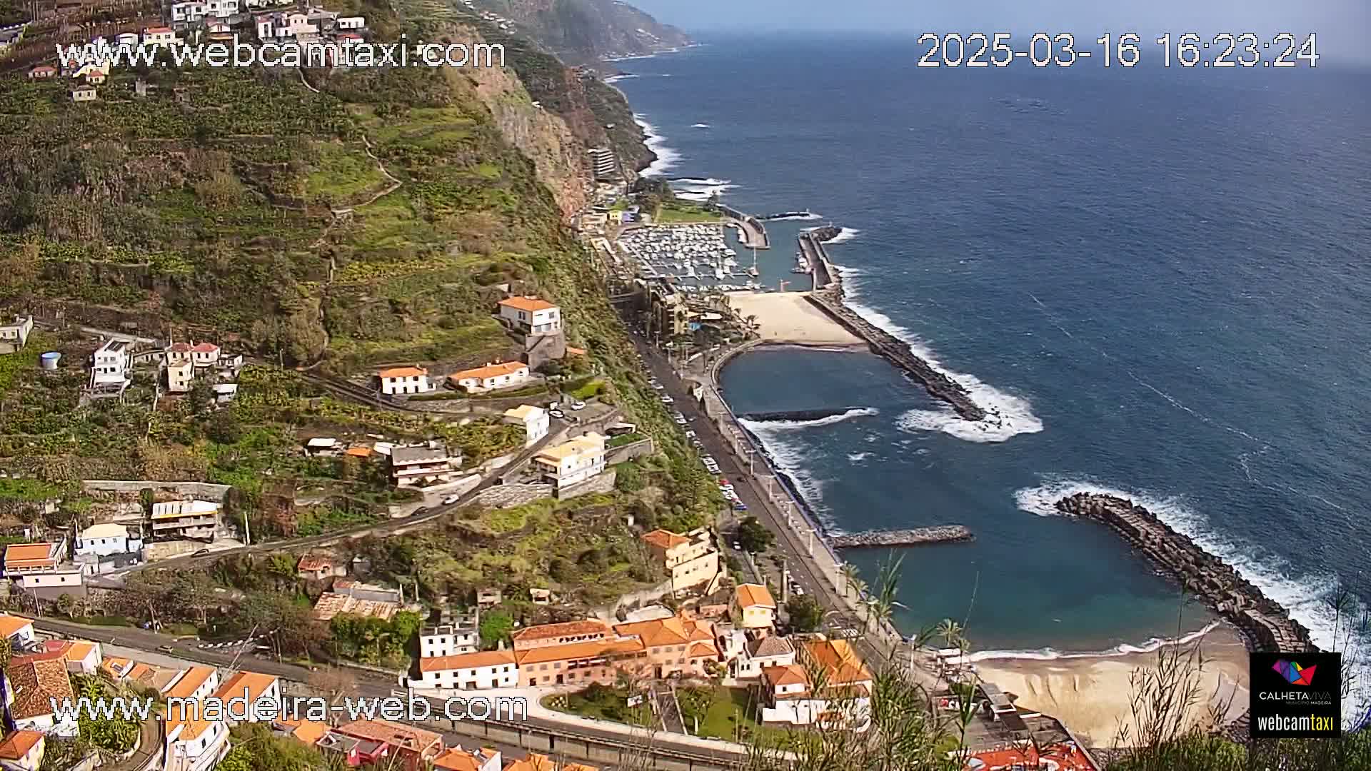 A coastal town built on a steep hillside overlooks a calm ocean with a small harbor and sandy beach on a sunny day.