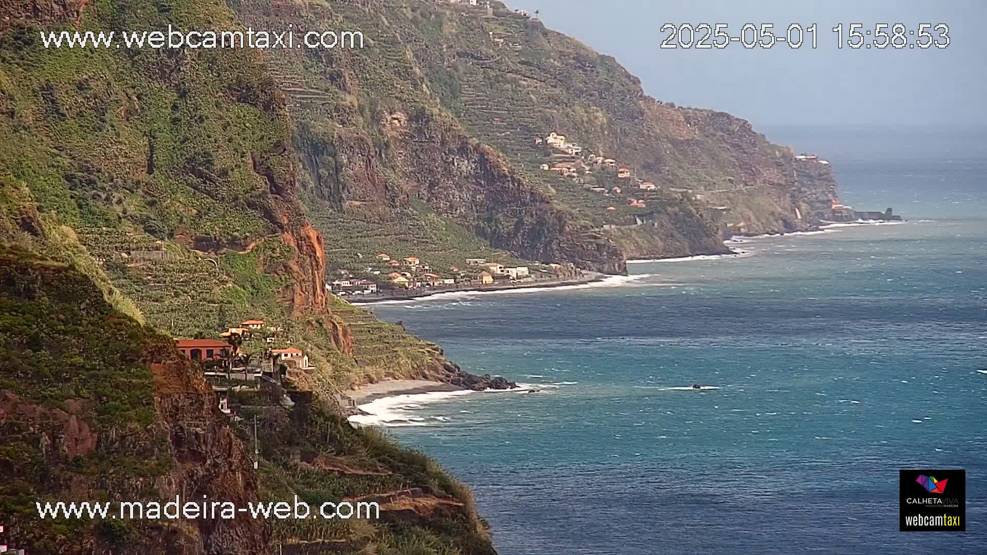 A coastal village nestled into a steep, verdant cliffside overlooks a dark-blue ocean under a mostly clear sky.