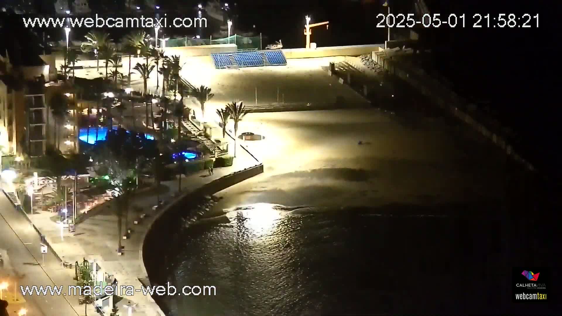 An illuminated sandy beach at night, next to a building with a pool, bordered by palm trees and a calm, dark ocean.