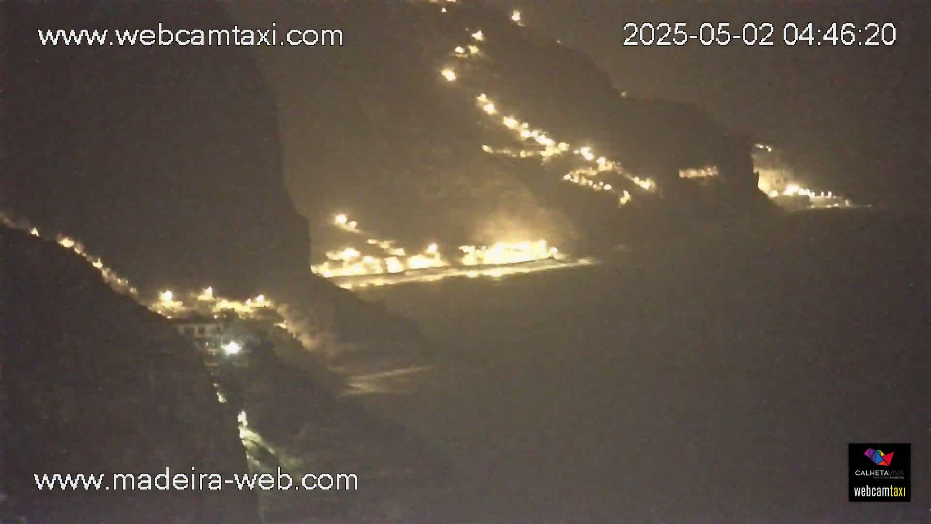 A nighttime, long shot shows a dark, rocky coastline with scattered lights along the cliffside and near the water's edge.
