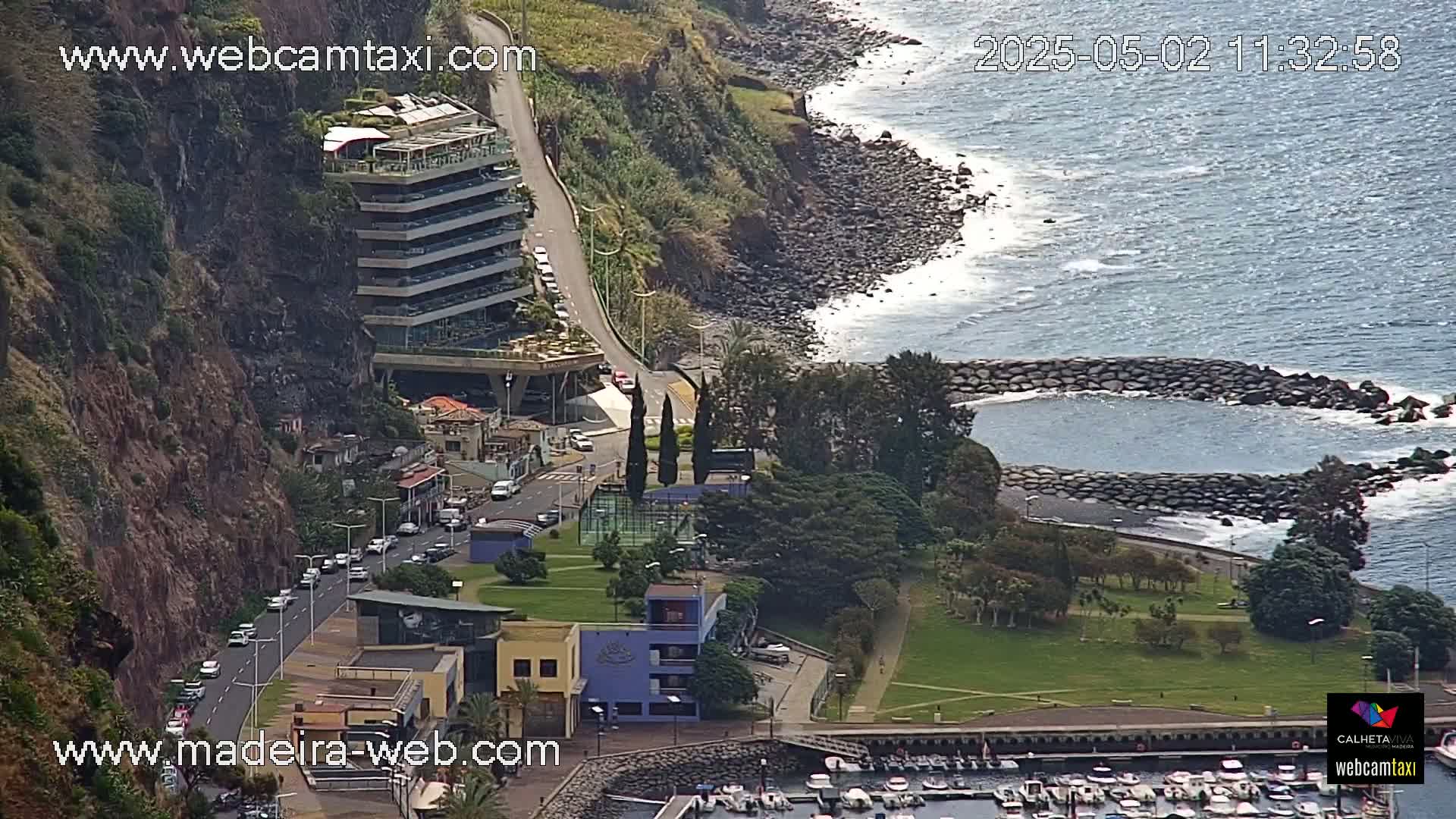 A coastal scene shows a multi-story building on a cliff overlooking a calm ocean with waves lapping a rocky shore, a small harbor filled with boats, and green grassy areas with trees.