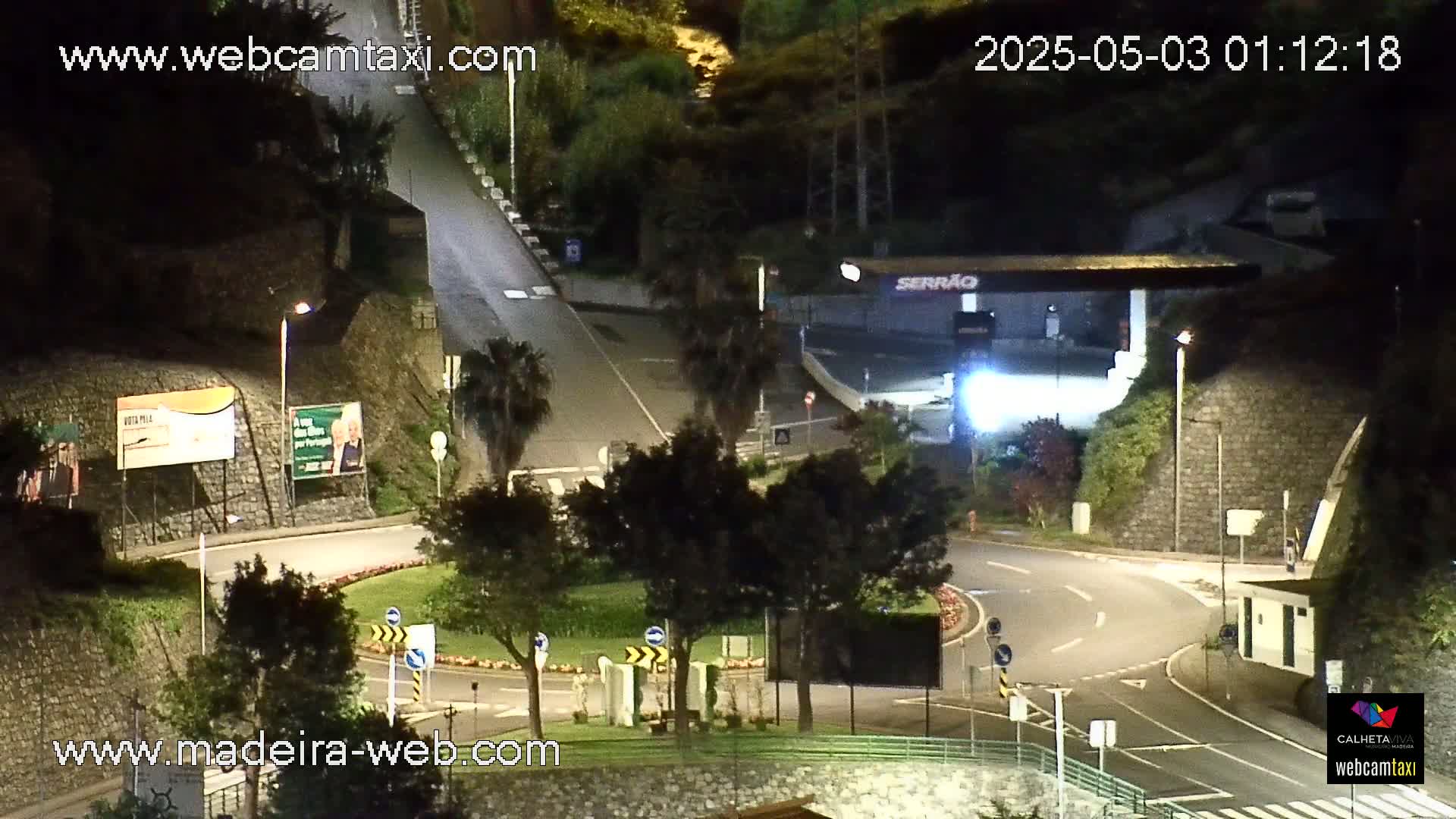 An aerial nighttime view shows a curving roadway with streetlights illuminating a roundabout surrounded by dark green trees and stone walls.
