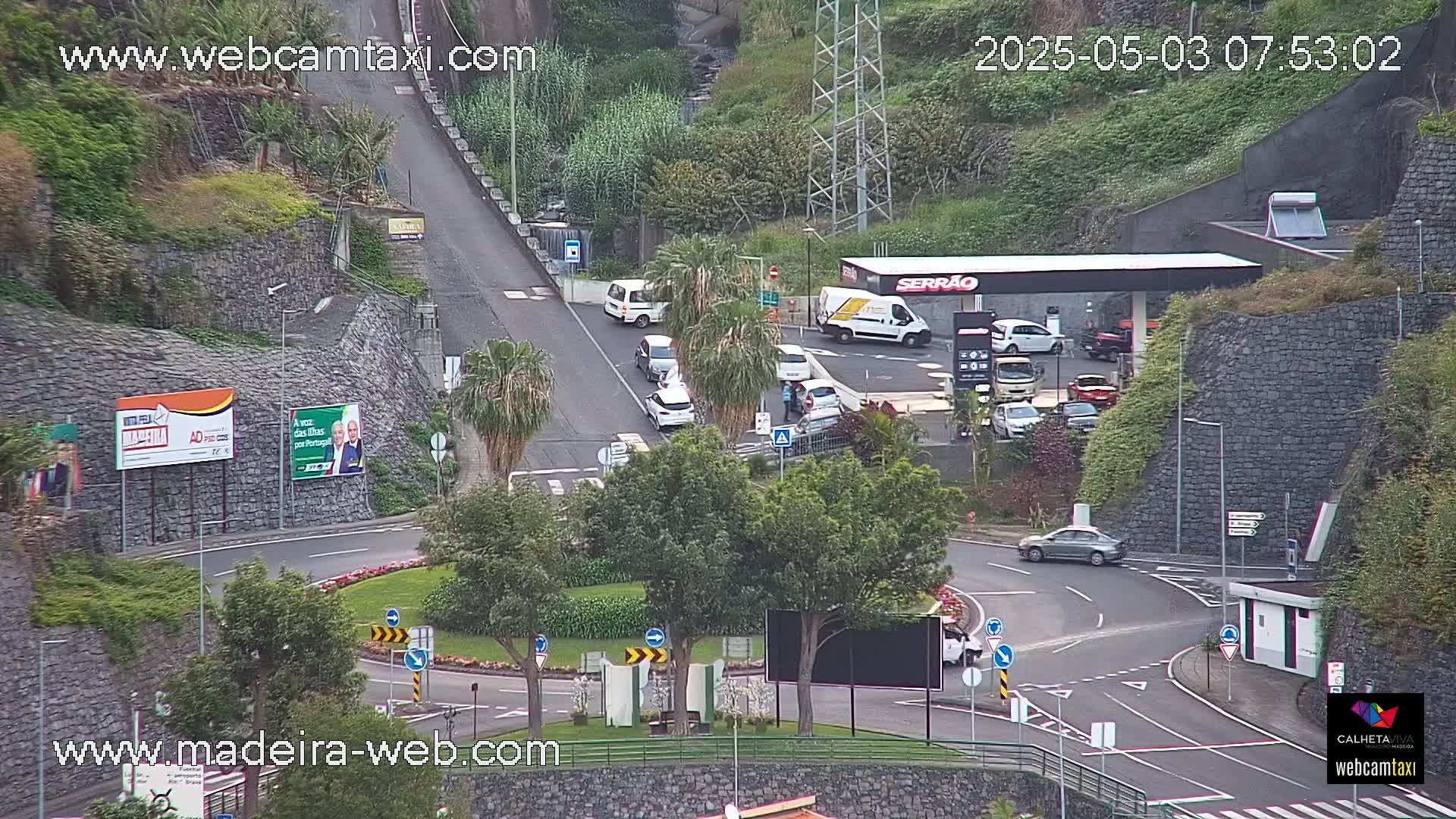 A gas station and roundabout are visible in a hilly, green area with several vehicles and trees, under partly cloudy conditions.