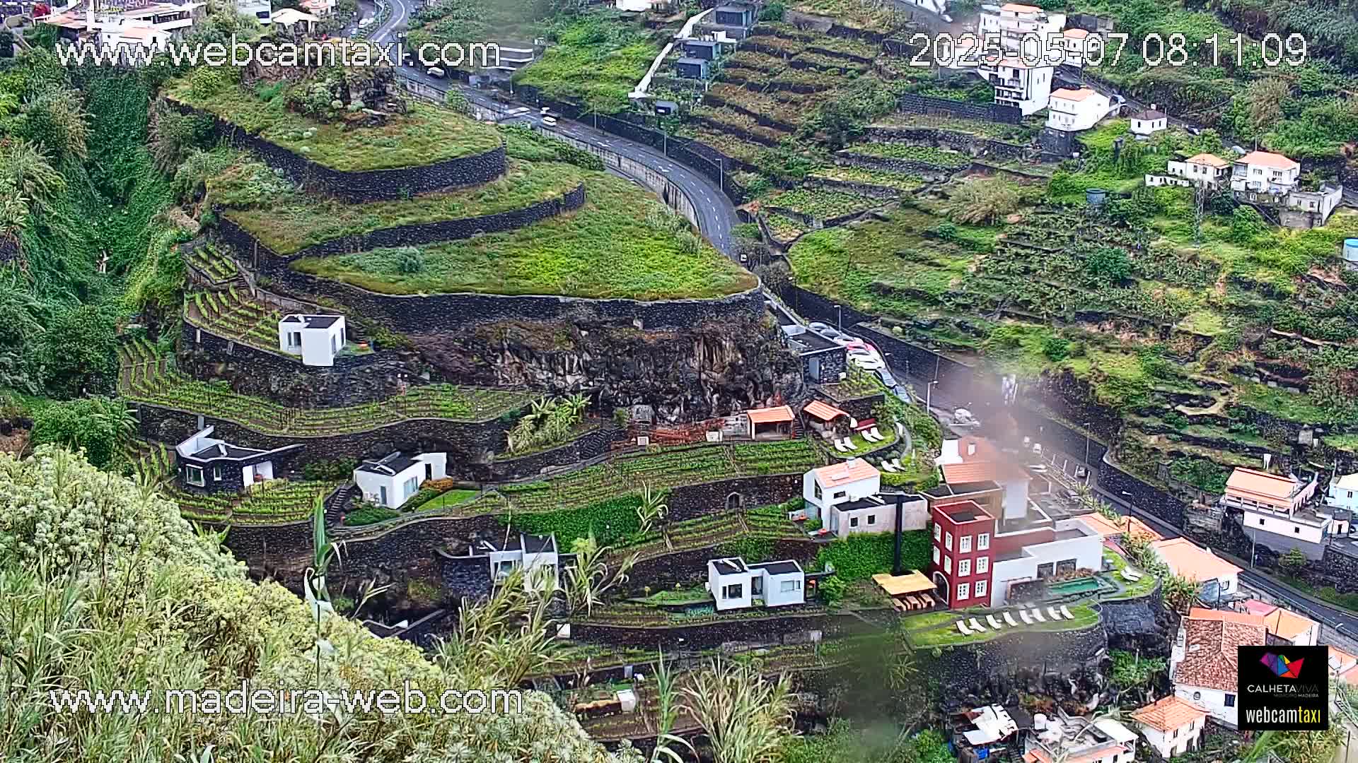 A hillside village with terraced gardens and modern houses is visible, viewed from above in rainy weather.