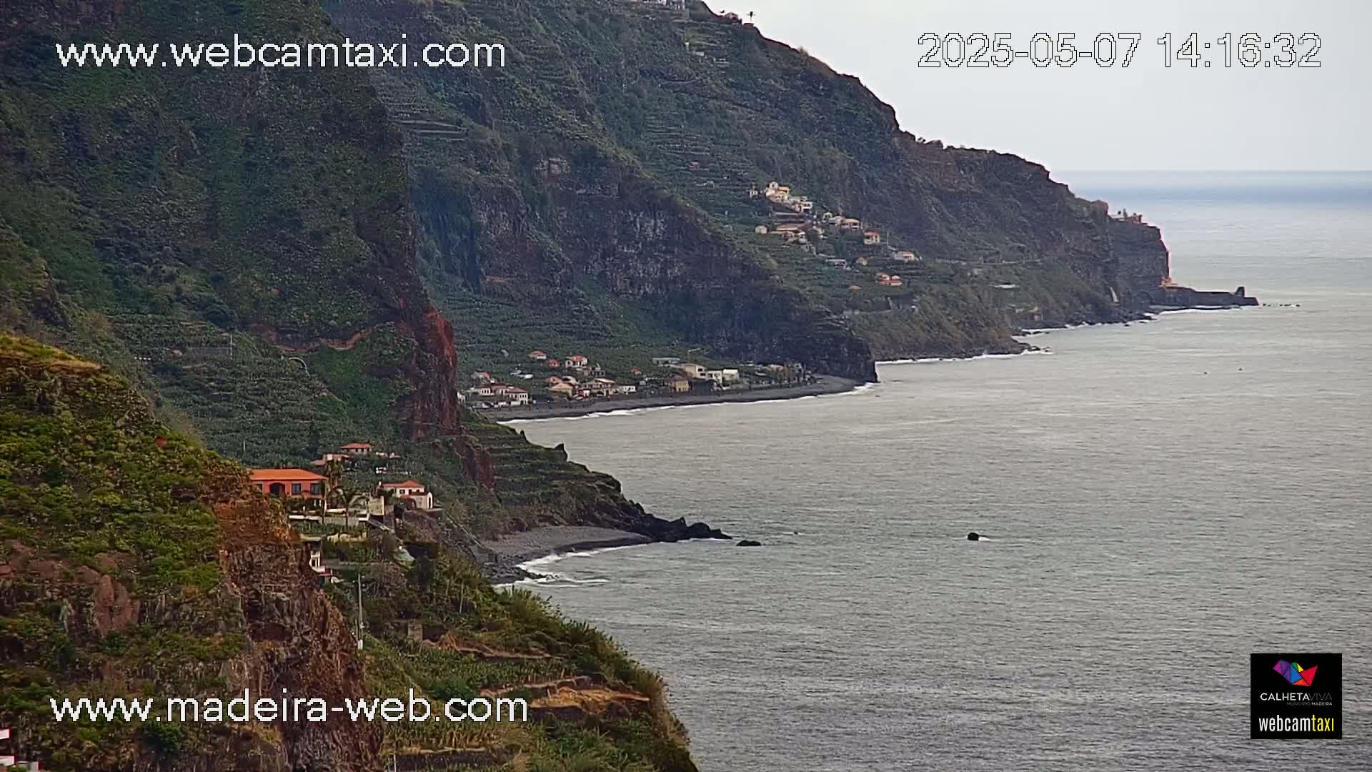 A dark, rocky coastline with a few buildings on the cliffs overlooks a calm, gray ocean under an overcast sky.