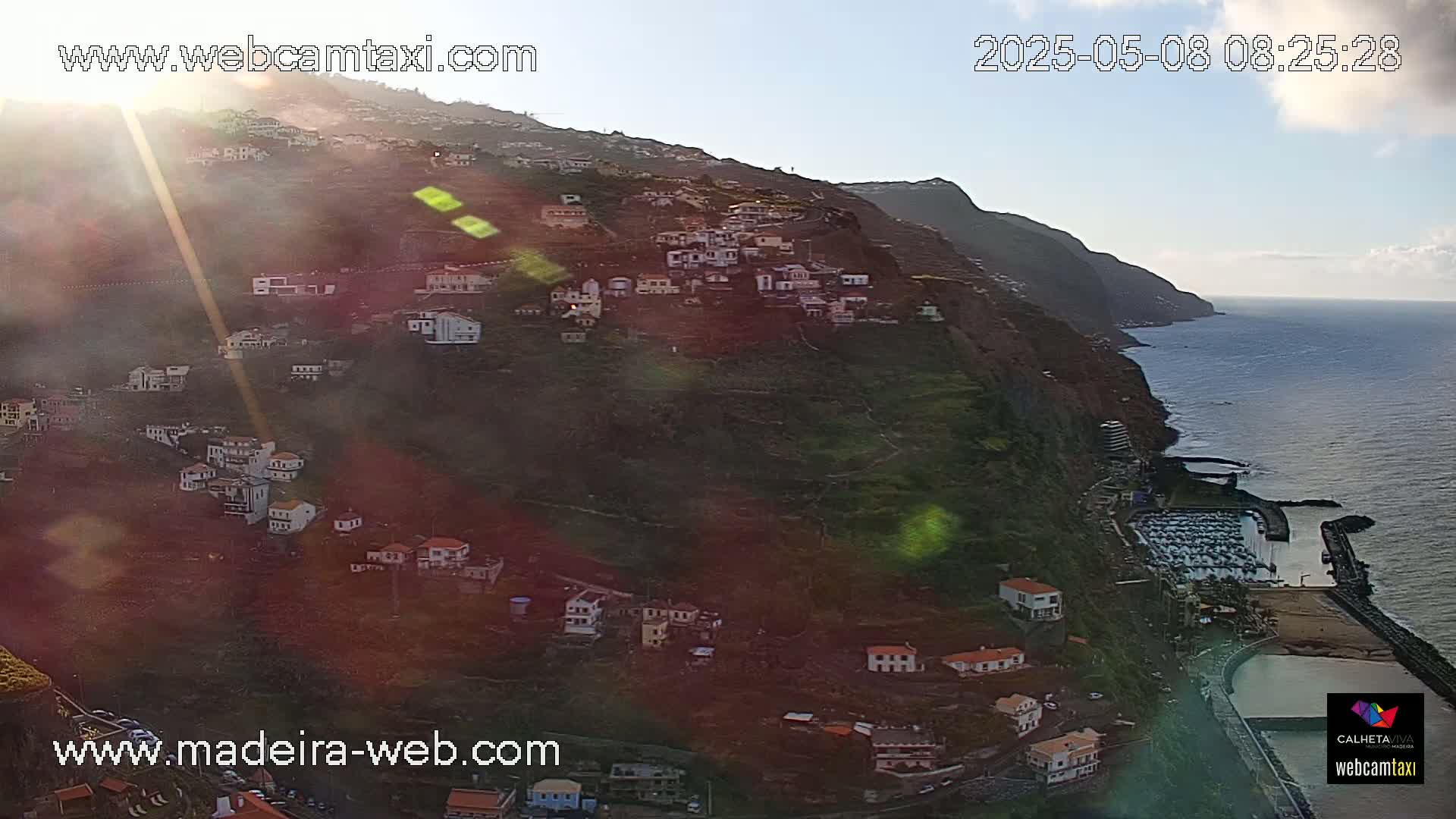 A sun-drenched hillside village clings to a steep cliff overlooking a calm ocean, with a marina visible below.