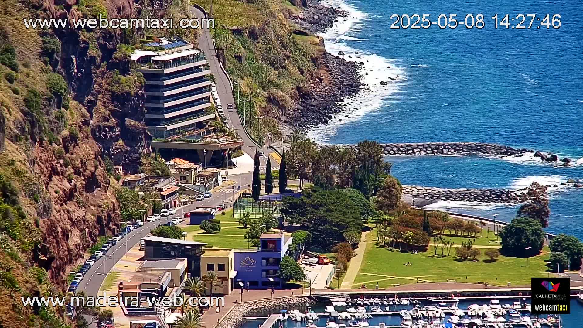 A coastal town with a marina, a cliffside hotel, and a park is seen on a sunny day, with a blue ocean and some waves breaking against the shore.