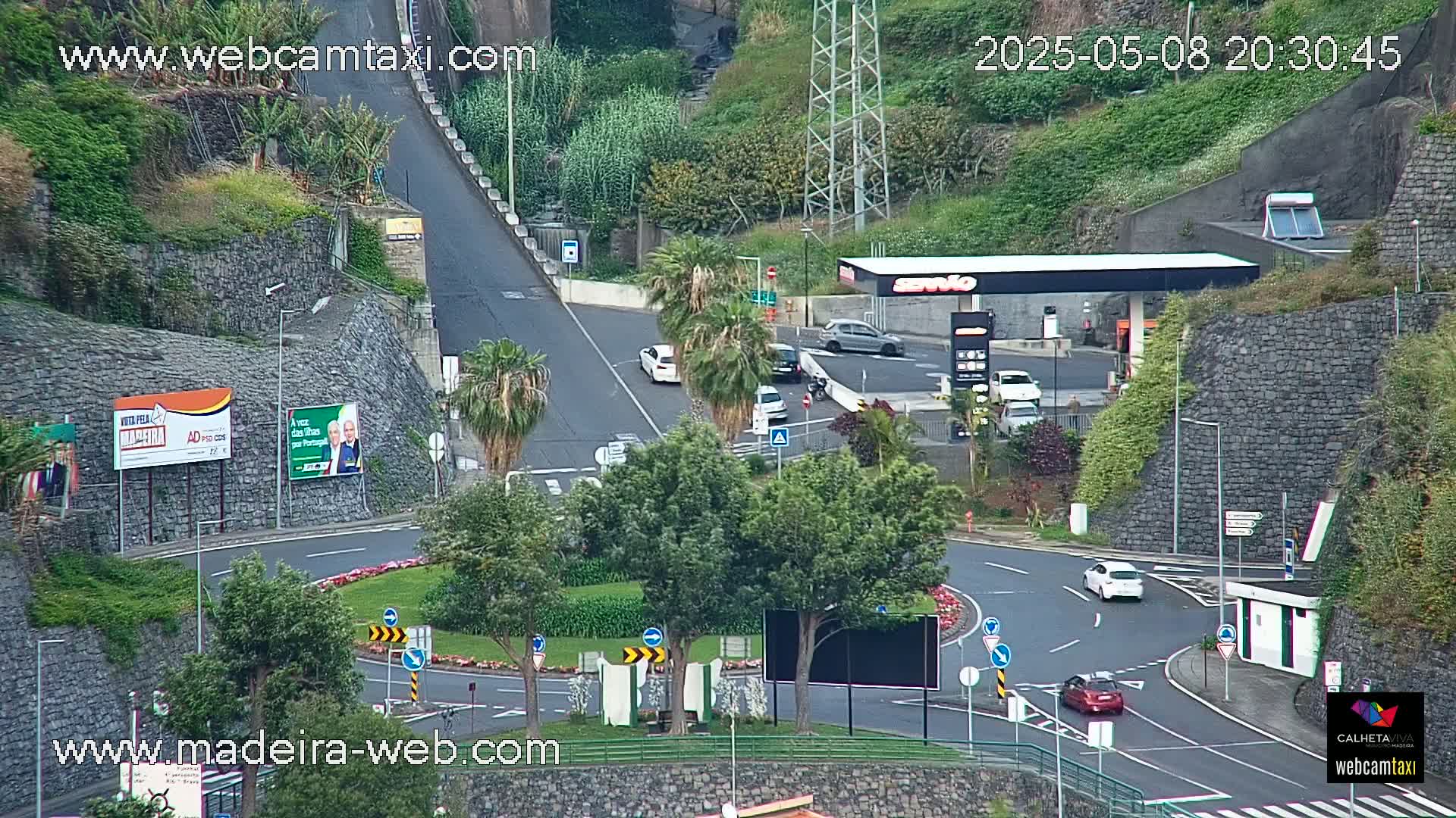 A roundabout with several cars and a gas station is situated on a hillside, overlooking a roadway that ascends steeply into the distance on an overcast day.