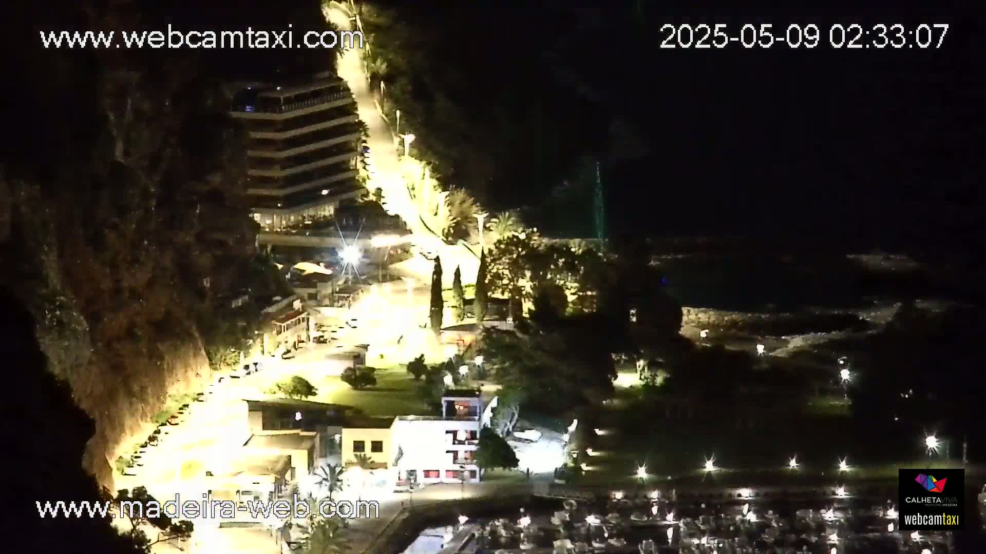 A nighttime aerial view shows a coastal town with illuminated buildings, roads, and a marina, nestled against a dark hillside.