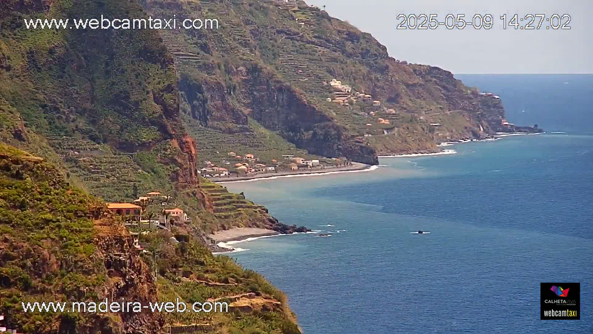 A coastal view shows a dark-colored beach nestled between a steep, terraced hillside and a calm, blue ocean under a clear sky.