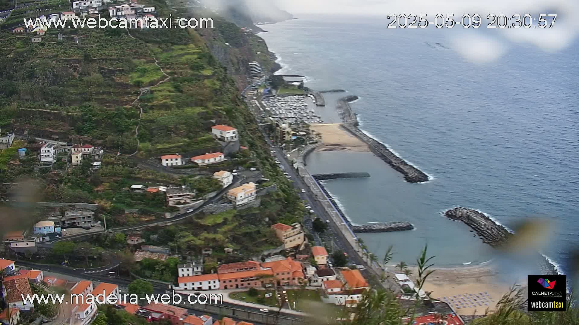 A coastal town nestled on a steep hillside overlooking a calm ocean with a marina and sandy beach, under an overcast sky.