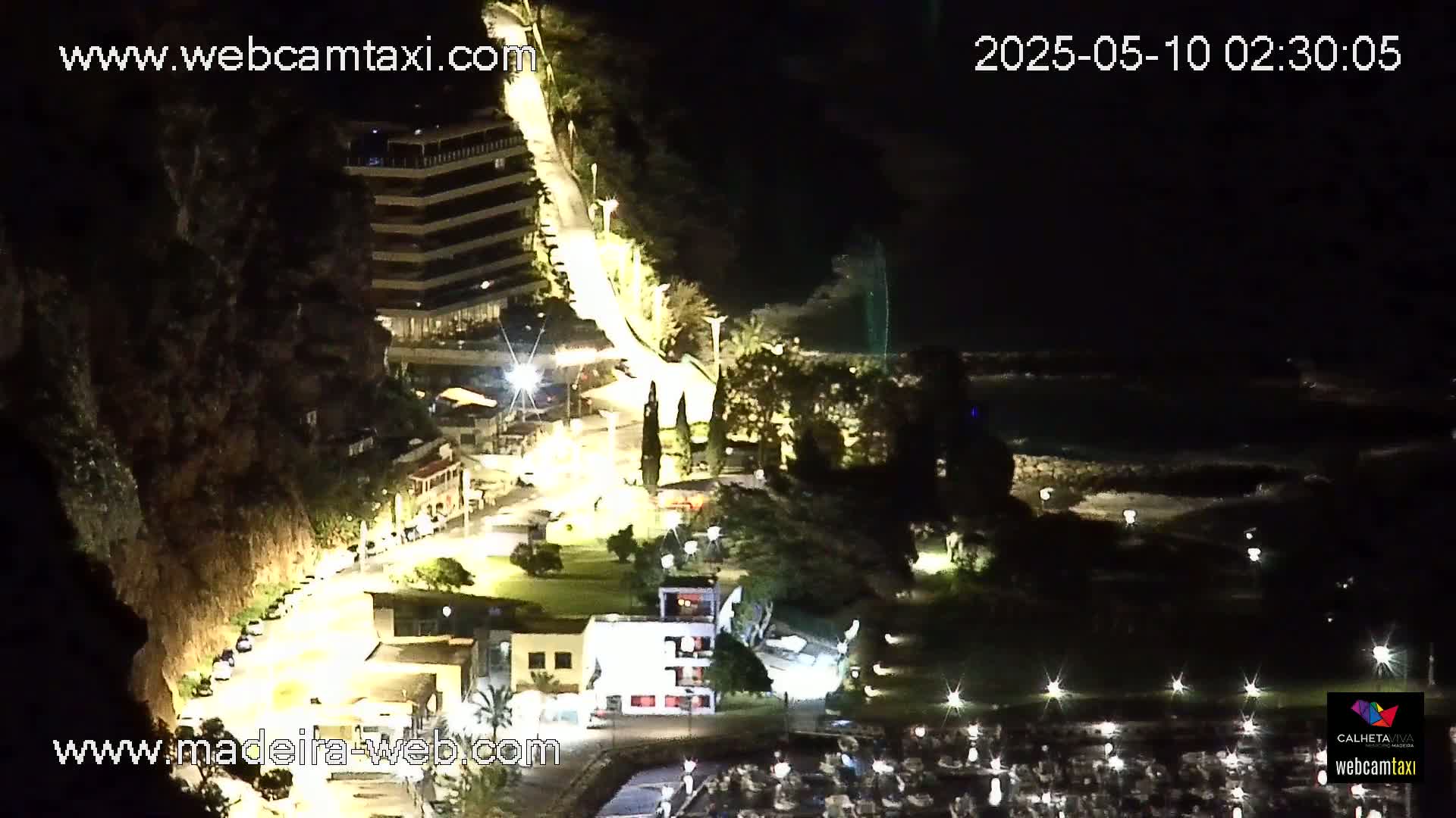A nighttime, overhead view of a coastal town with a curving road illuminated by streetlights, buildings, and a marina.