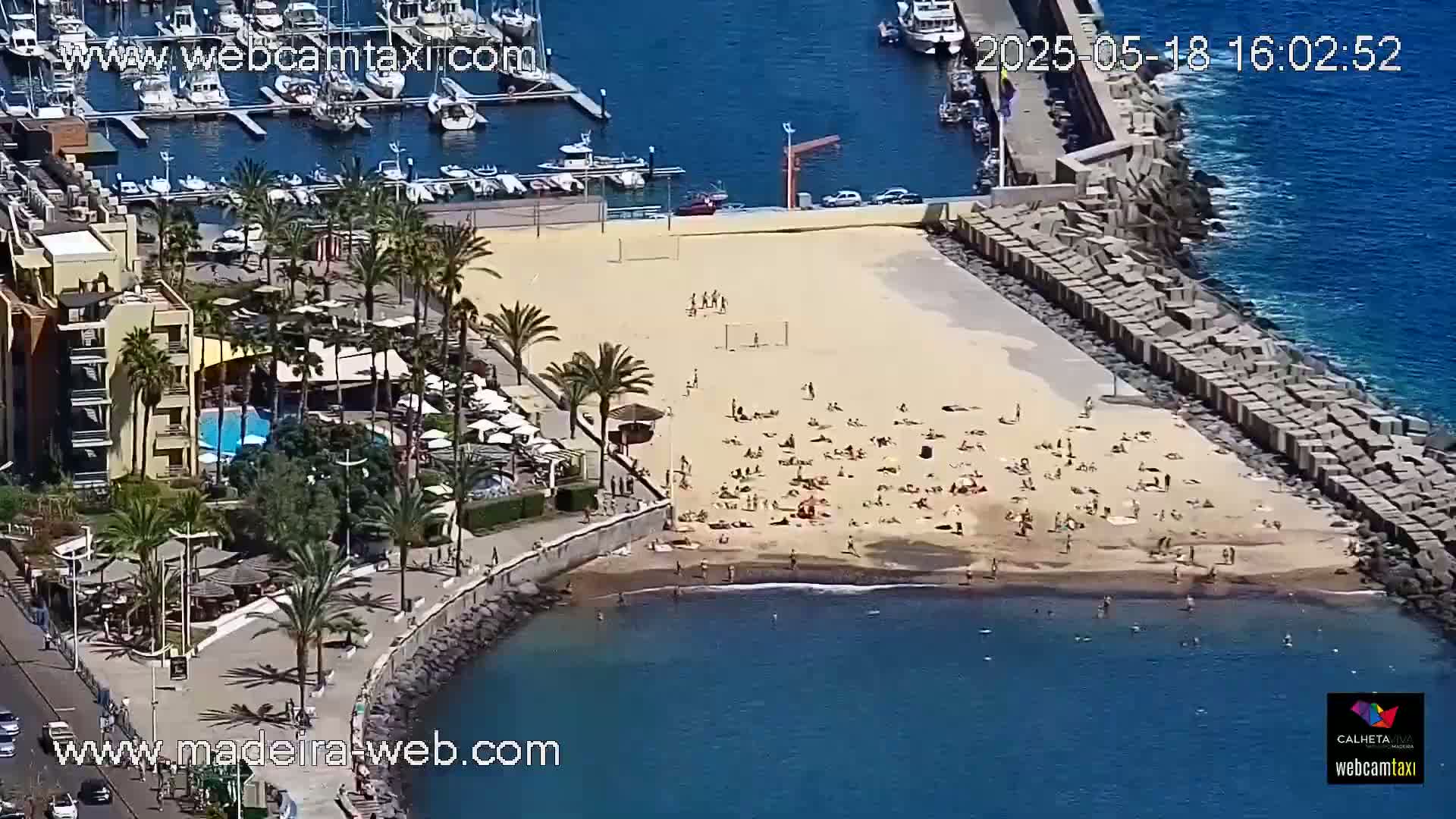 An aerial view shows a sandy beach with many people sunbathing, adjacent to a marina filled with boats, a resort with a pool, and a rocky breakwater extending into a calm, blue ocean under a sunny sky.